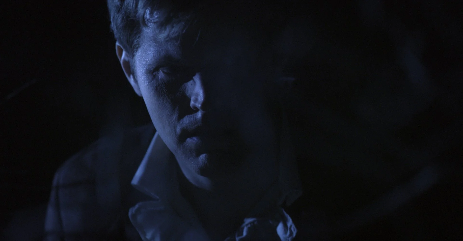 Dark, moody photo of a man's face partially illuminated in blue light, with shadows obscuring details.