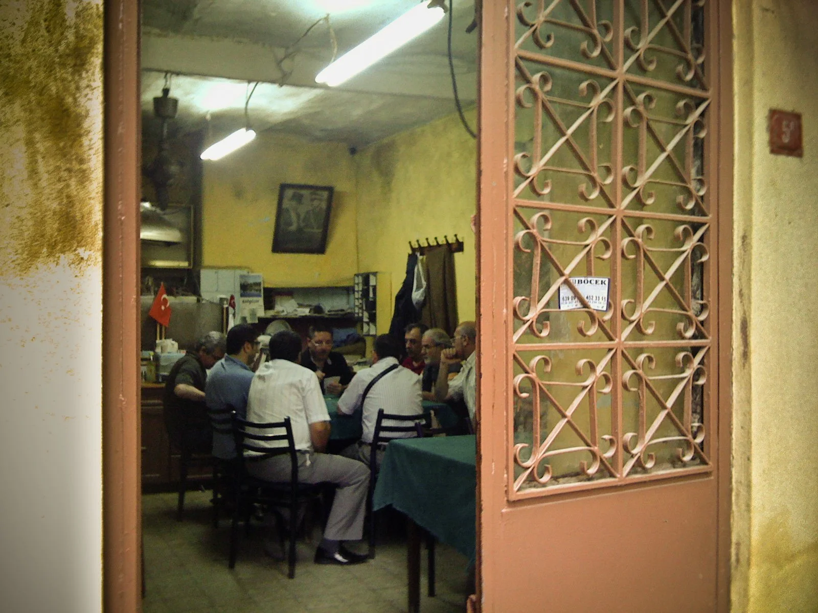 People sitting at tables inside a small restaurant, seen through an open door with decorative ironwork. The interior has yellow walls with a framed picture, fluorescent ceiling lights, and kitchen shelves with dishes and appliances.