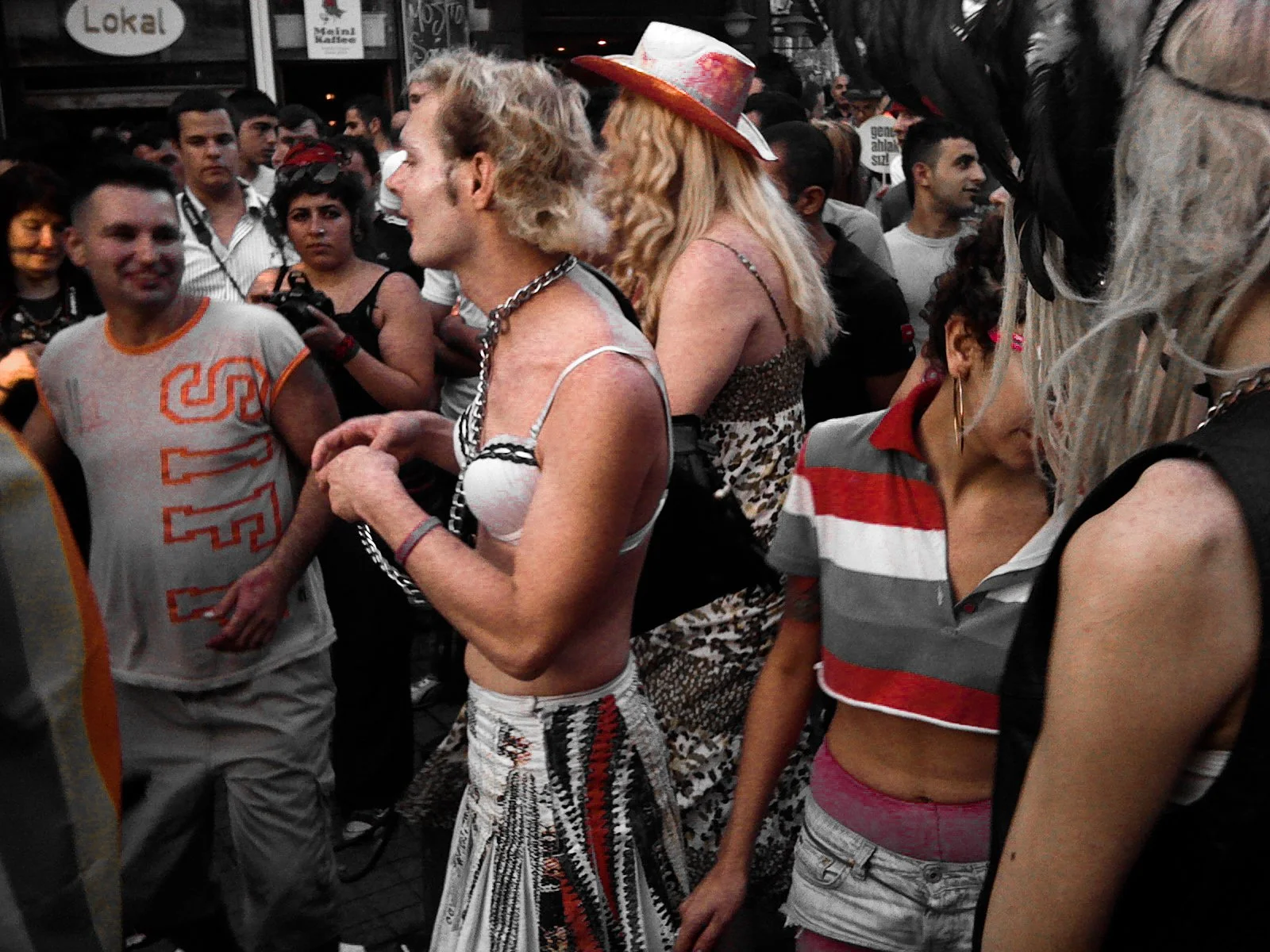 Group of people gathered outdoors, socializing, with some wearing casual and retro-style clothing, one woman with long blond hair and a hat in the center, and others of various ages and styles in the crowd.