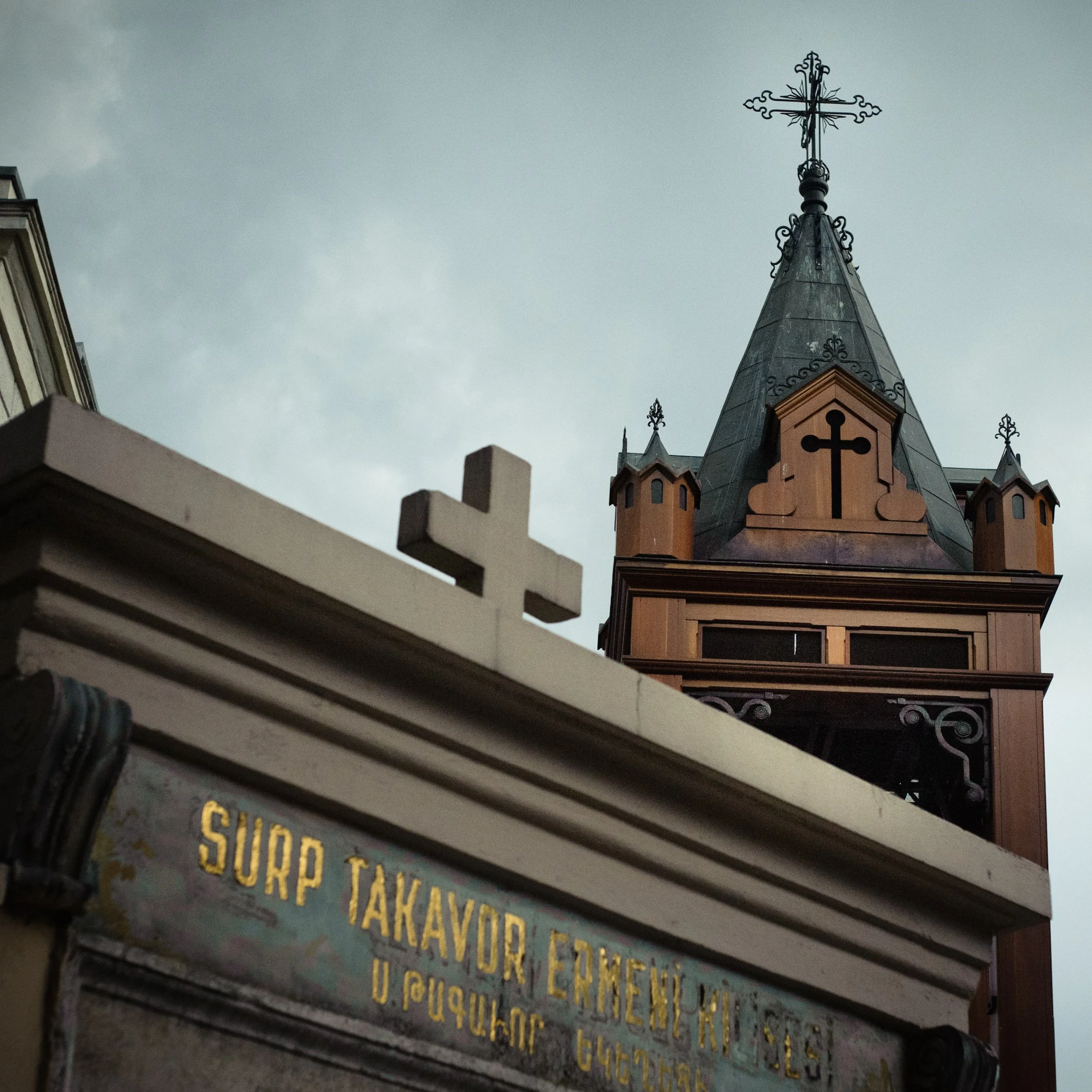 A close-up of a building with a sign reading "SURP TAKAVOR ENEUKI NELS" in gold lettering, and a white cross in the foreground. In the background, there is a tall, ornate church steeple topped with a cross and a detailed wrought iron cross at the ver