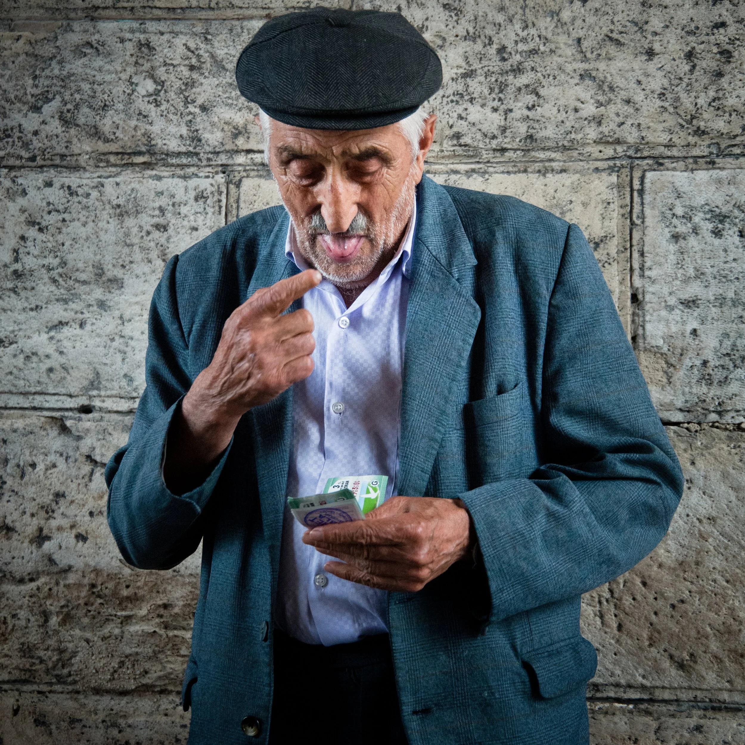 An elderly man with gray hair and a beard, wearing a black flat cap, a blue blazer, and a white shirt, stands against a stone wall. He is looking at a handful of money, pointing to his face with his right finger.