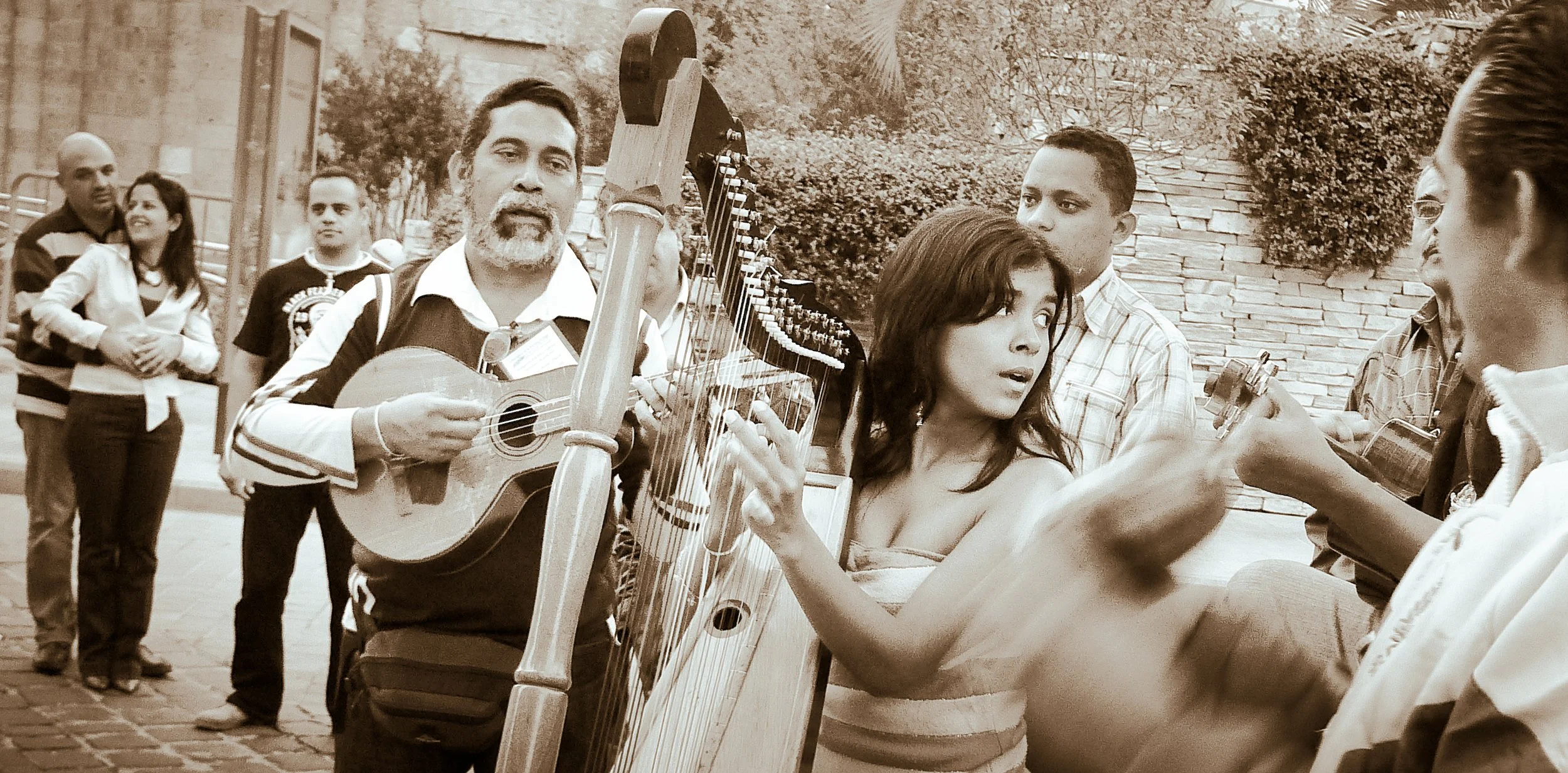 A group of street performers playing guitar and harp for a small crowd, including a woman singing, engaging with passersby.