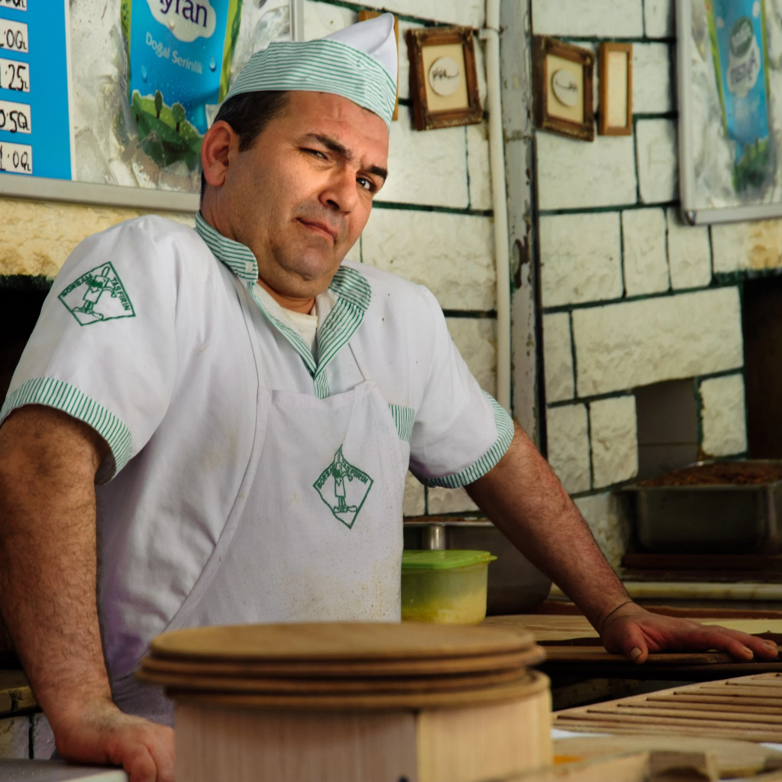 A man in a white chef's uniform and hat standing behind a counter in a kitchen, looking at the camera with a slight frown.