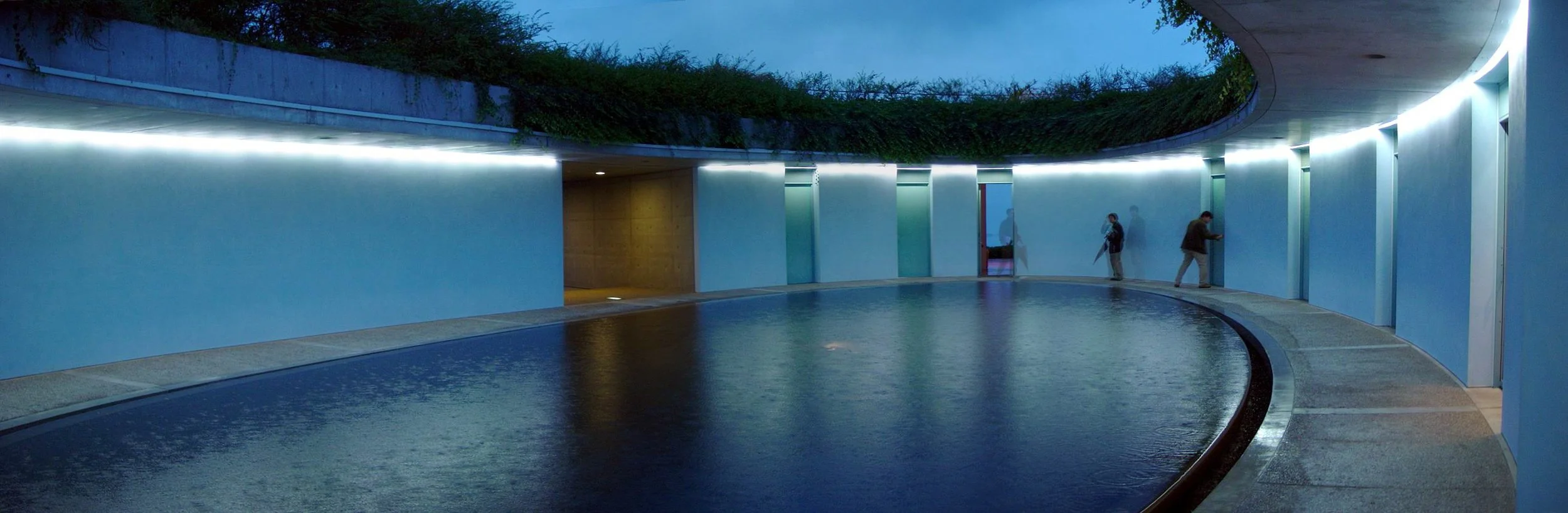 Modern building entrance with curved glass walls, outdoor water feature, and people walking near elevators.