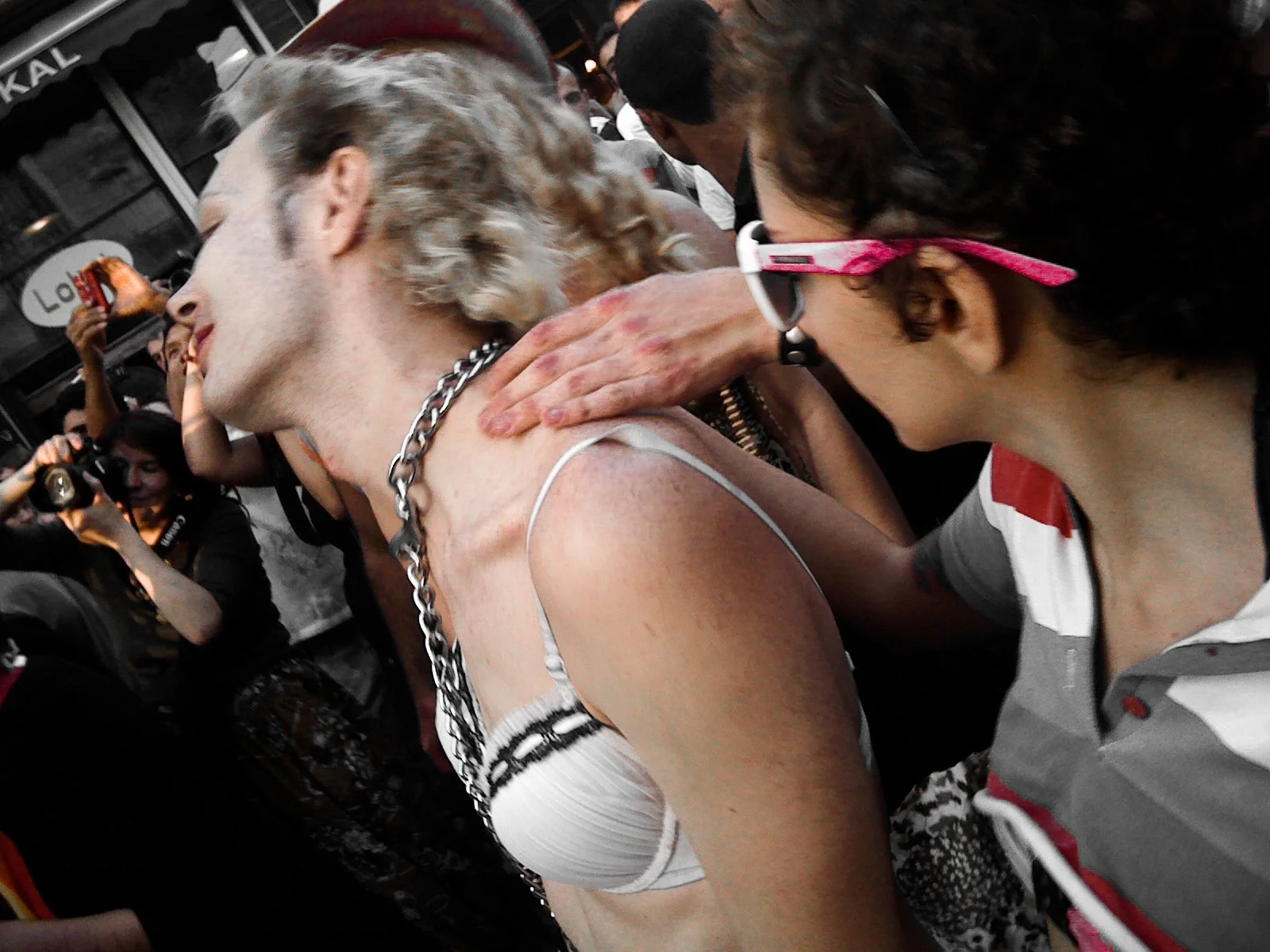 Person with short, curly hair receiving a neck massage from a woman wearing glasses at a crowded event.