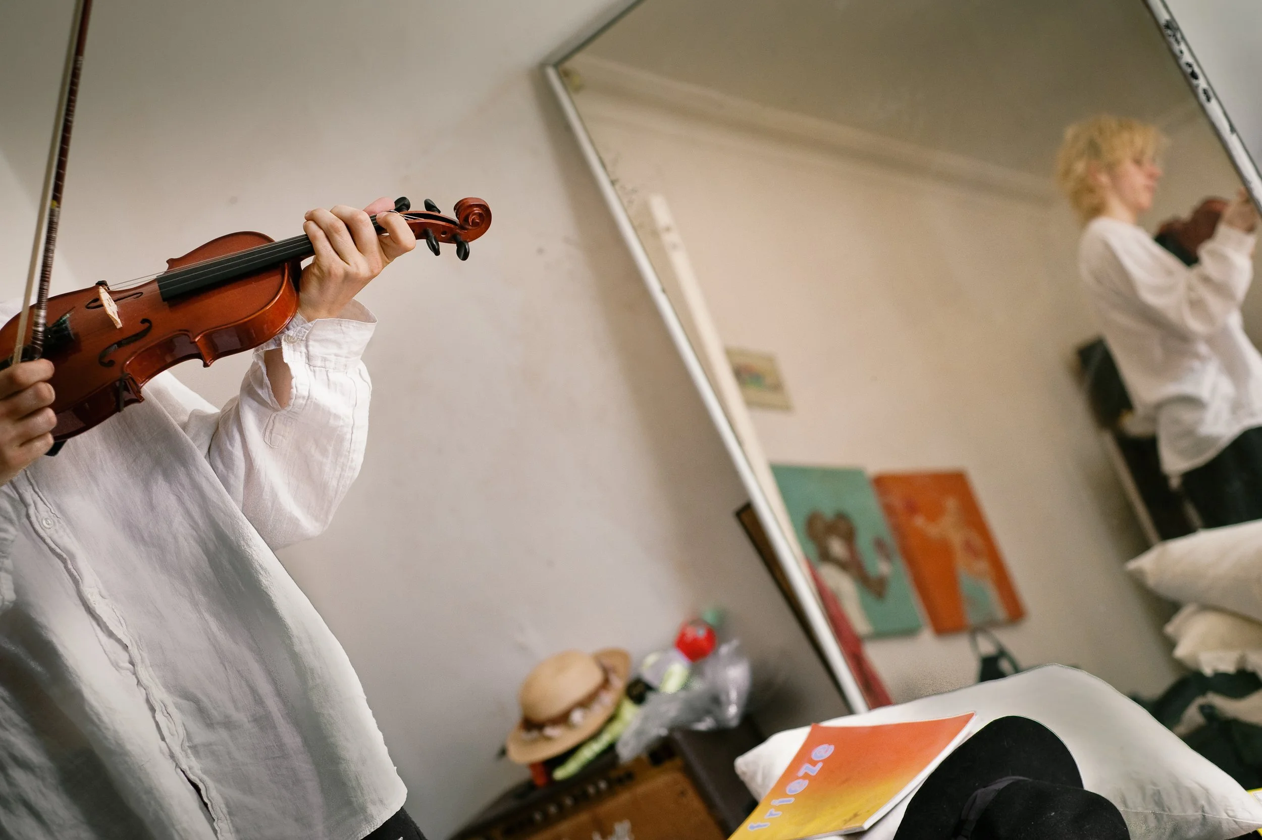 Person playing a violin in front of a mirror in a bedroom or hotel room, with artwork and a hamburger on a table visible in the background.
