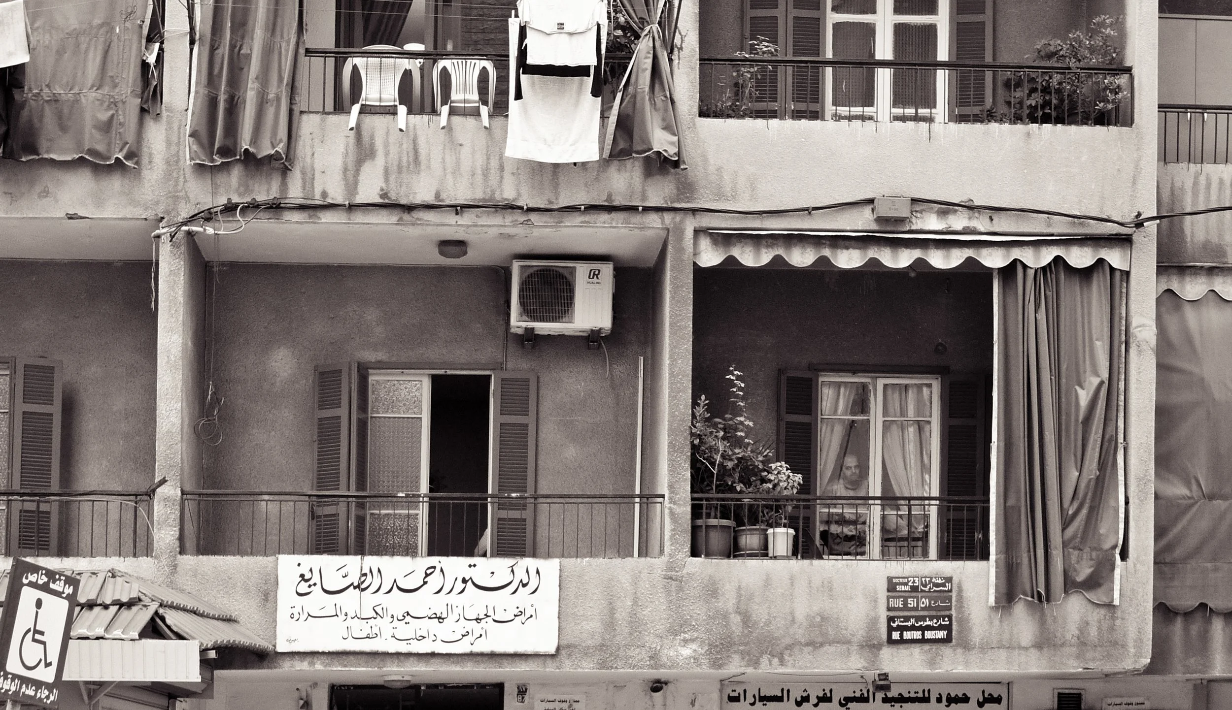 Apartment building facade with balconies, an air conditioning unit, curtains, nobody visible, and signs in Arabic at street level.