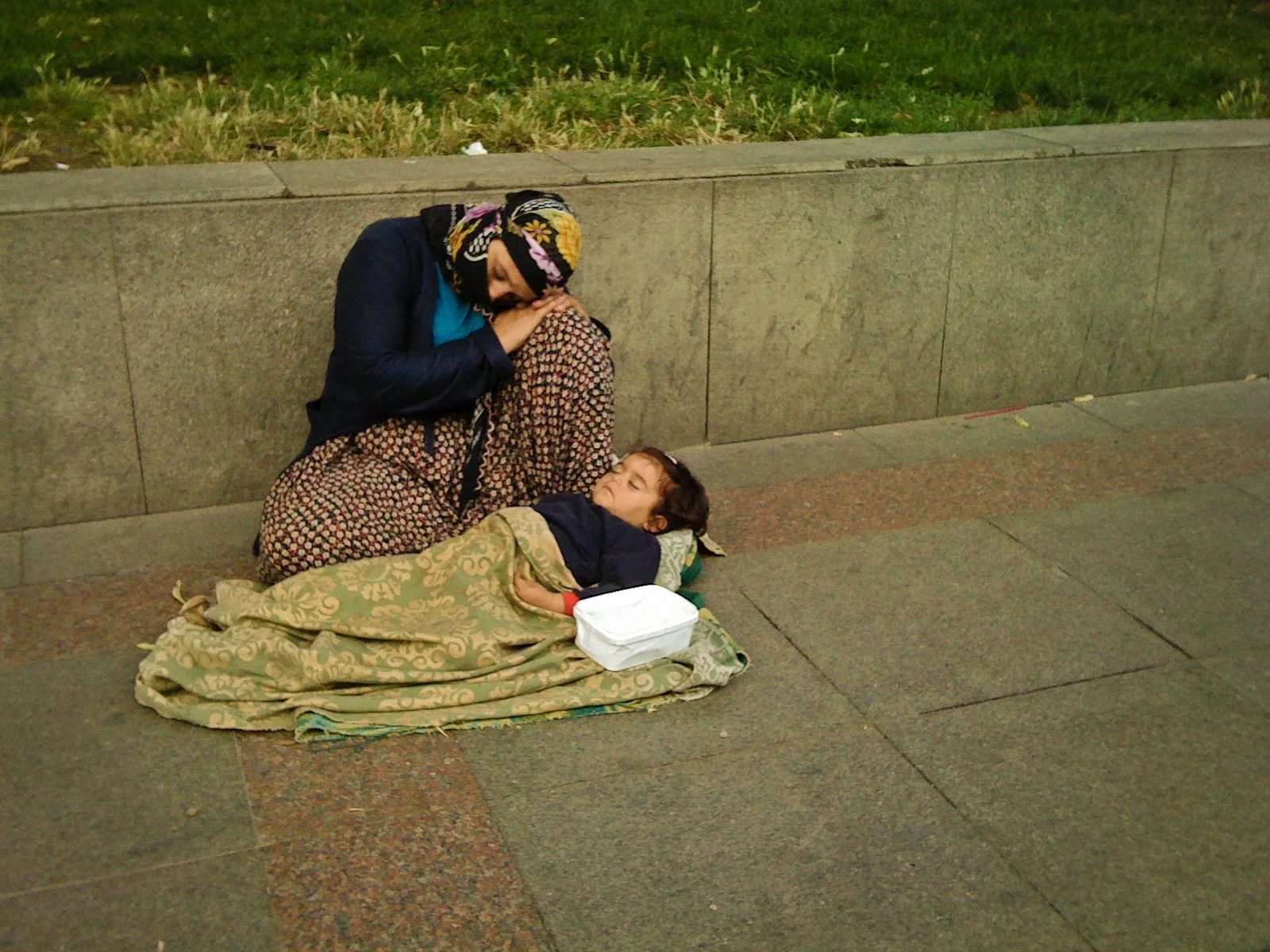 A woman in a dark jacket and colorful headscarf comforts a sleeping child lying on the sidewalk, wrapped in a blanket, next to a white food container.