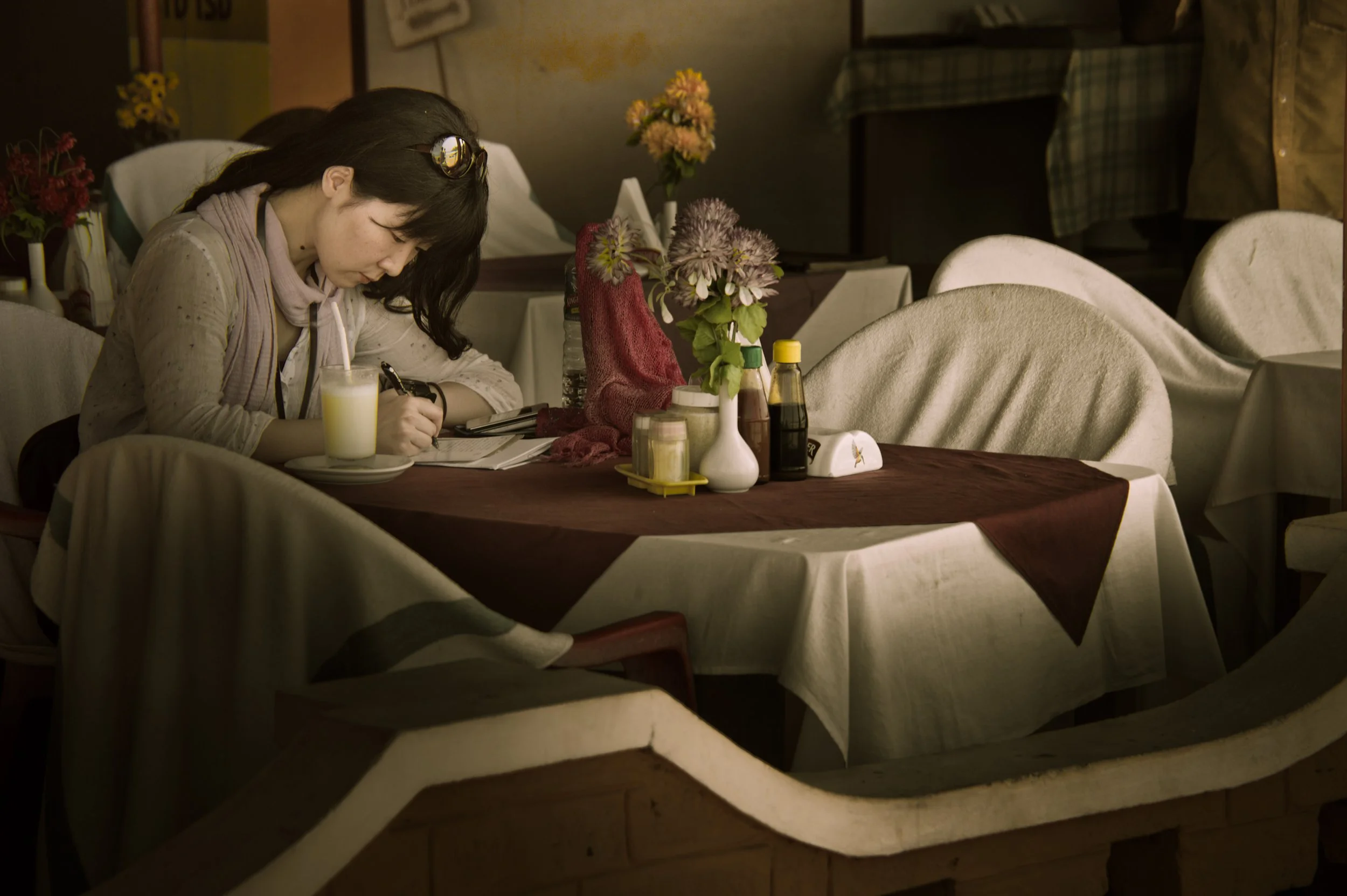 A woman sitting alone at a restaurant table, writing in a notebook, with a glass of juice nearby. The table is decorated with flowers and condiment bottles.
