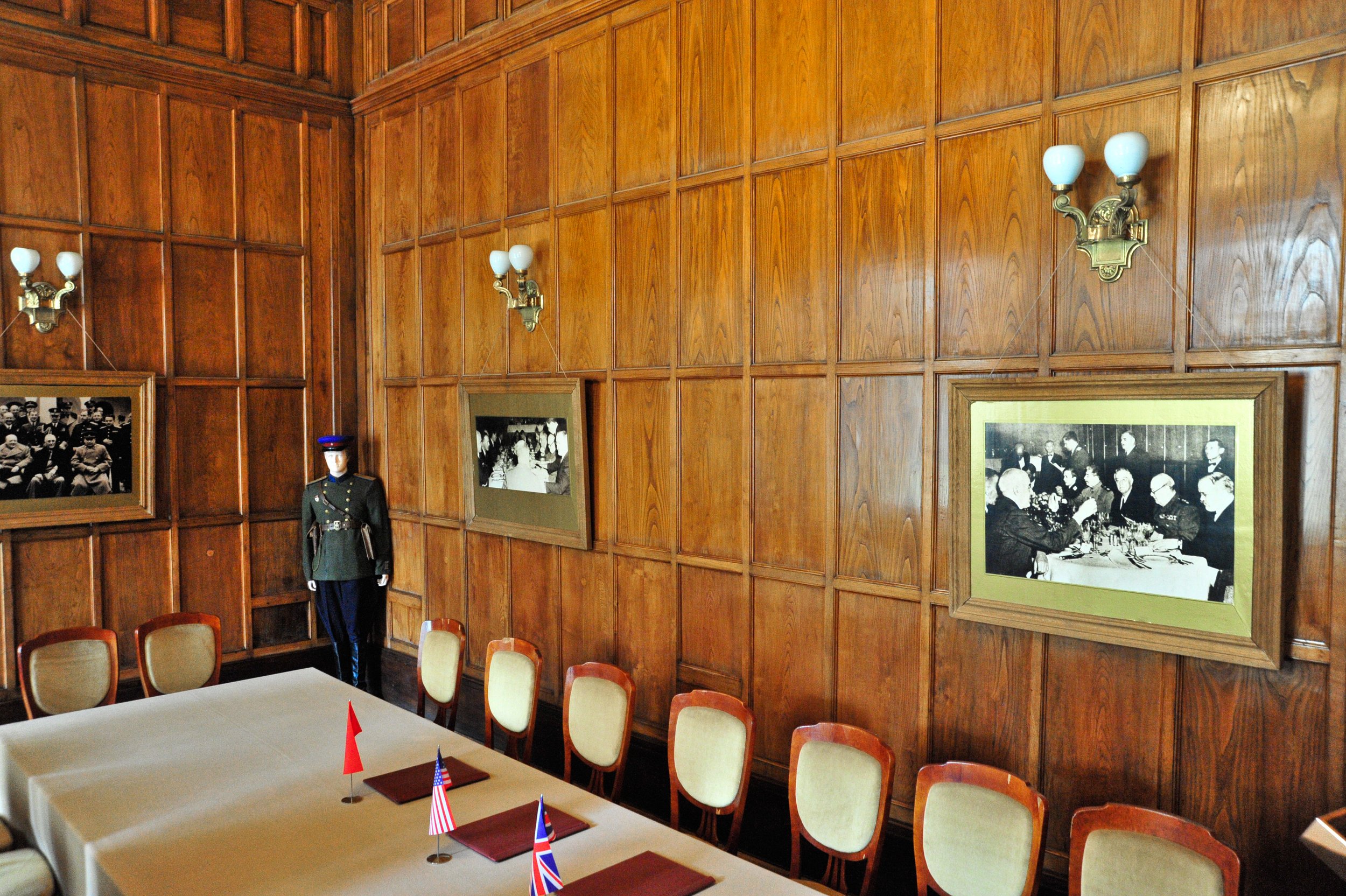 A formal dining room with a long table, six chairs, and three small flags of China, the United States, and the United Kingdom on the table. The room has wood-paneled walls, vintage black-and-white photographs in wooden frames, and wall-mounted light 