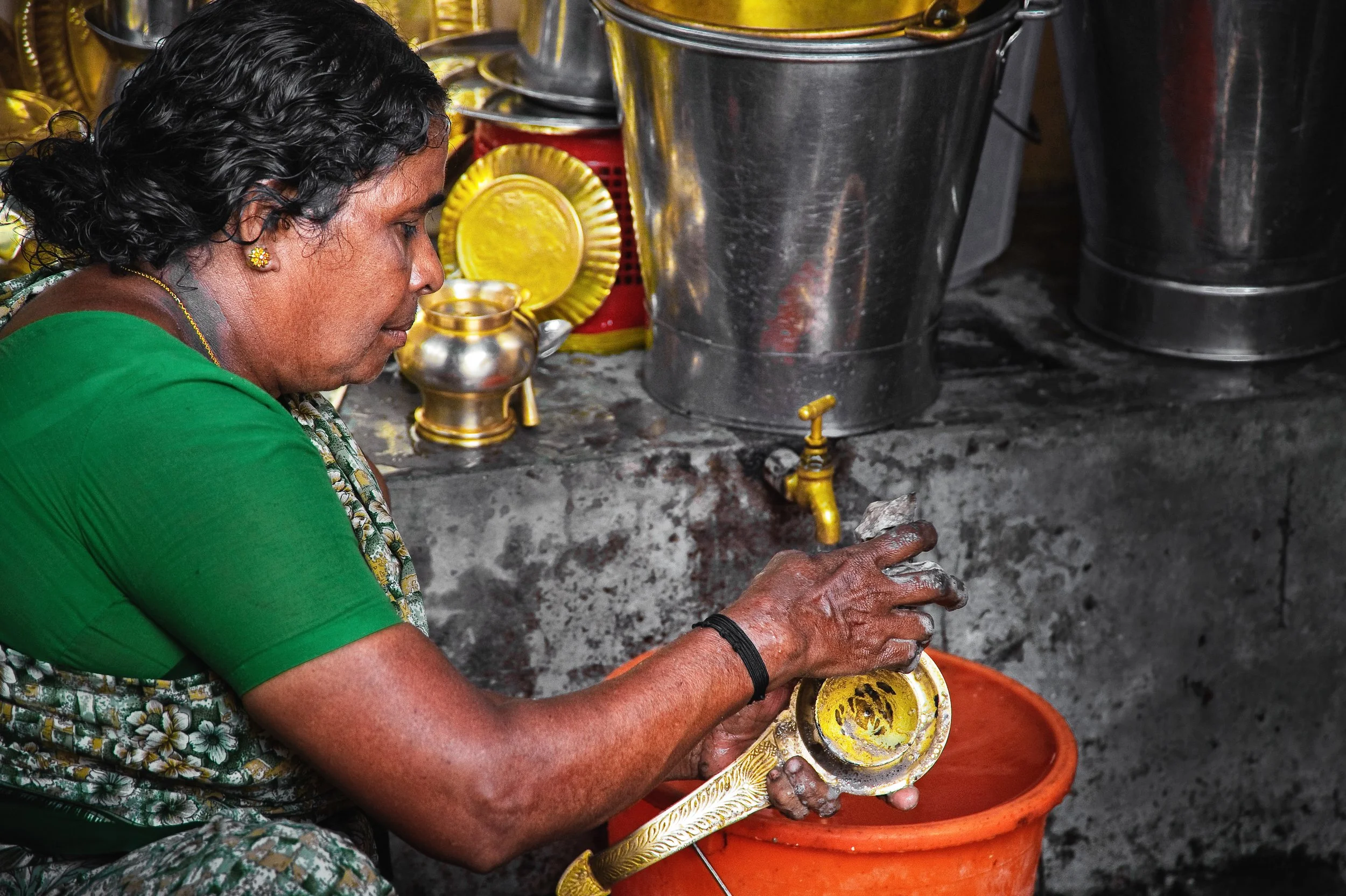 A woman with dark curly hair and gold earrings is washing a brass vessel in an orange bucket. She is wearing a green blouse with a floral saree and is working in a kitchen with metallic utensils and yellow metal dishes around her.