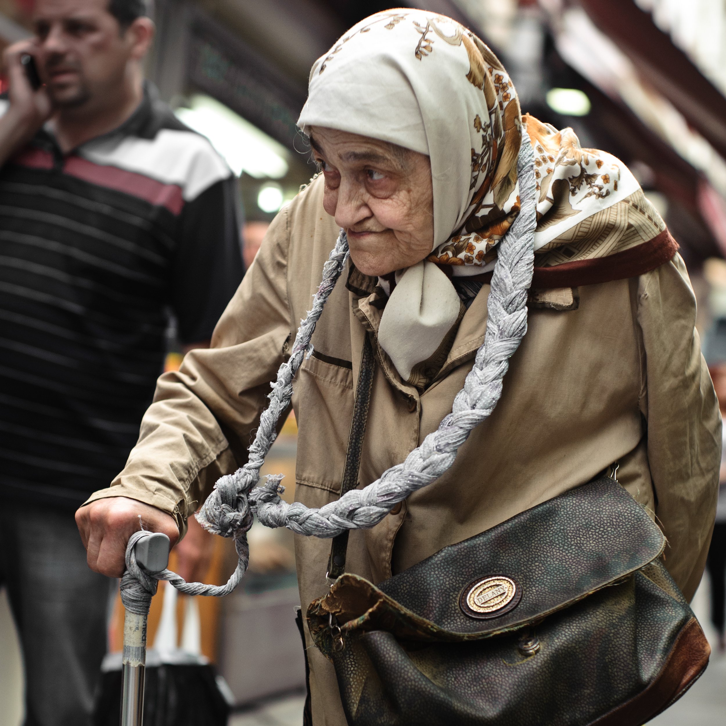 An elderly woman with a headscarf and beige coat using a cane, carrying a patterned shoulder bag, in a busy indoor market or shopping area.