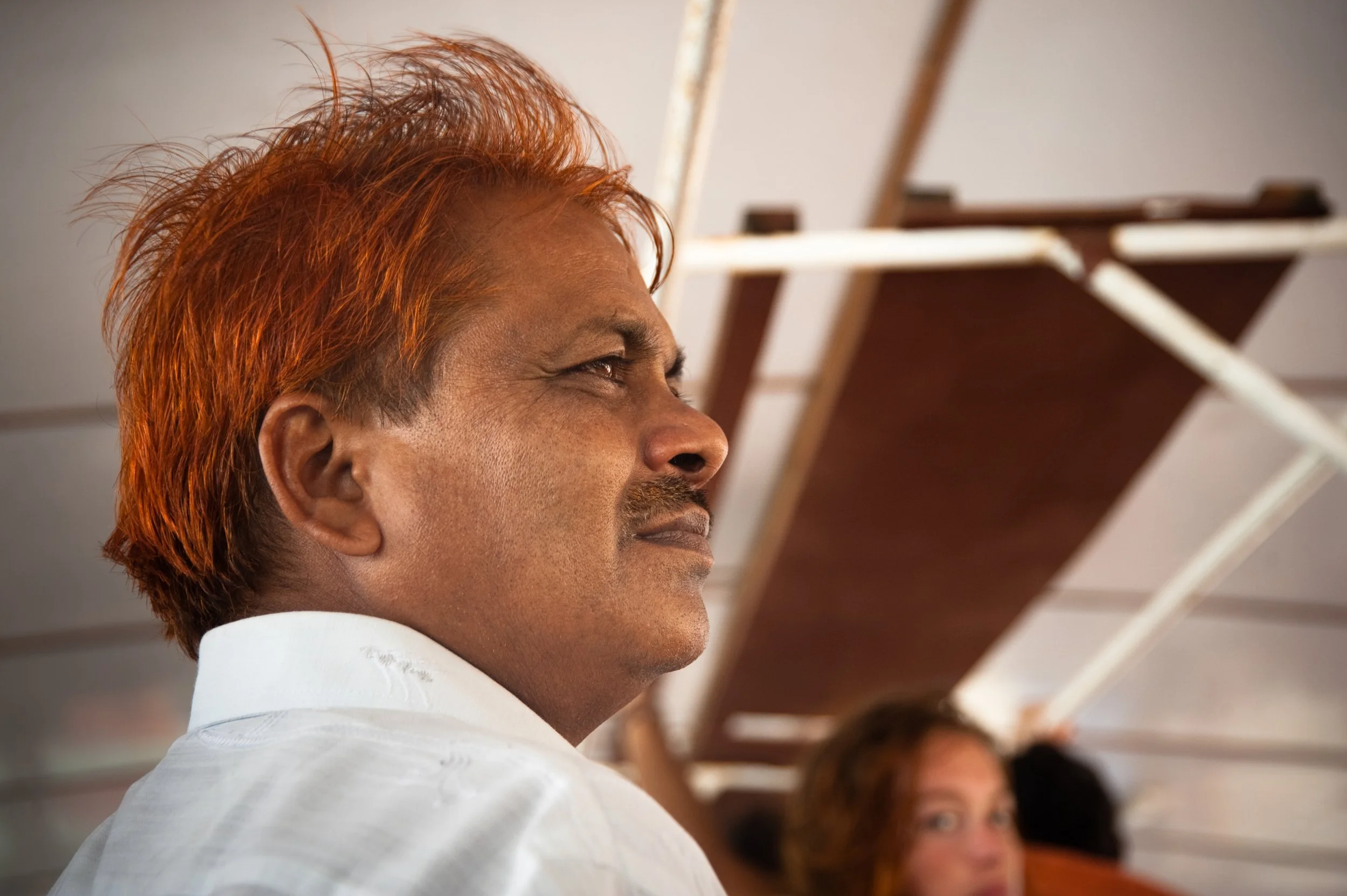 A man with reddish hair and a mustache, wearing a white shirt, sitting on a bus or train, with other passengers visible in the background.