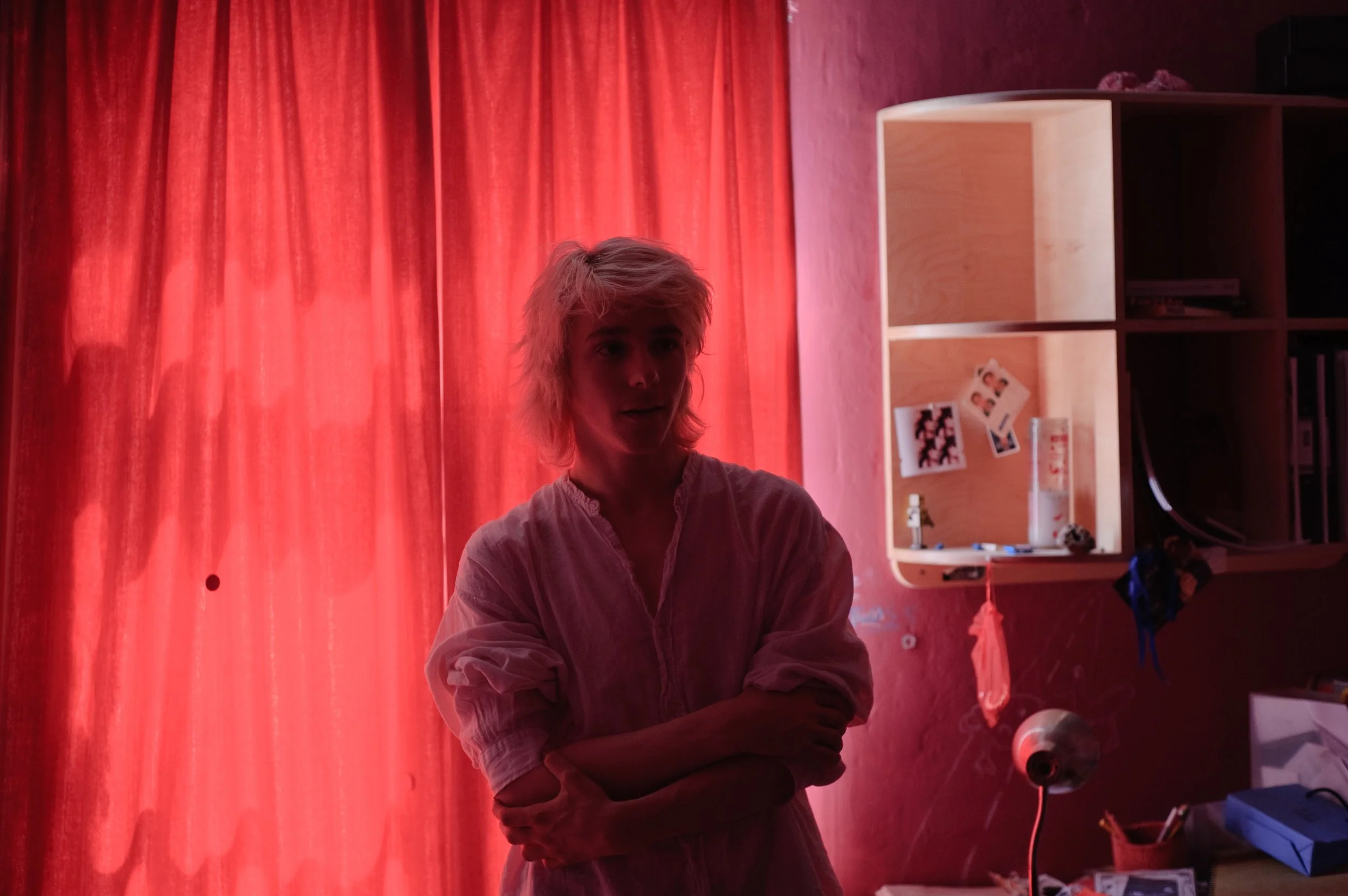 A person with light-colored, messy hair standing in a room with red curtains, with a bookshelf on the right side that has photos and various items on it.