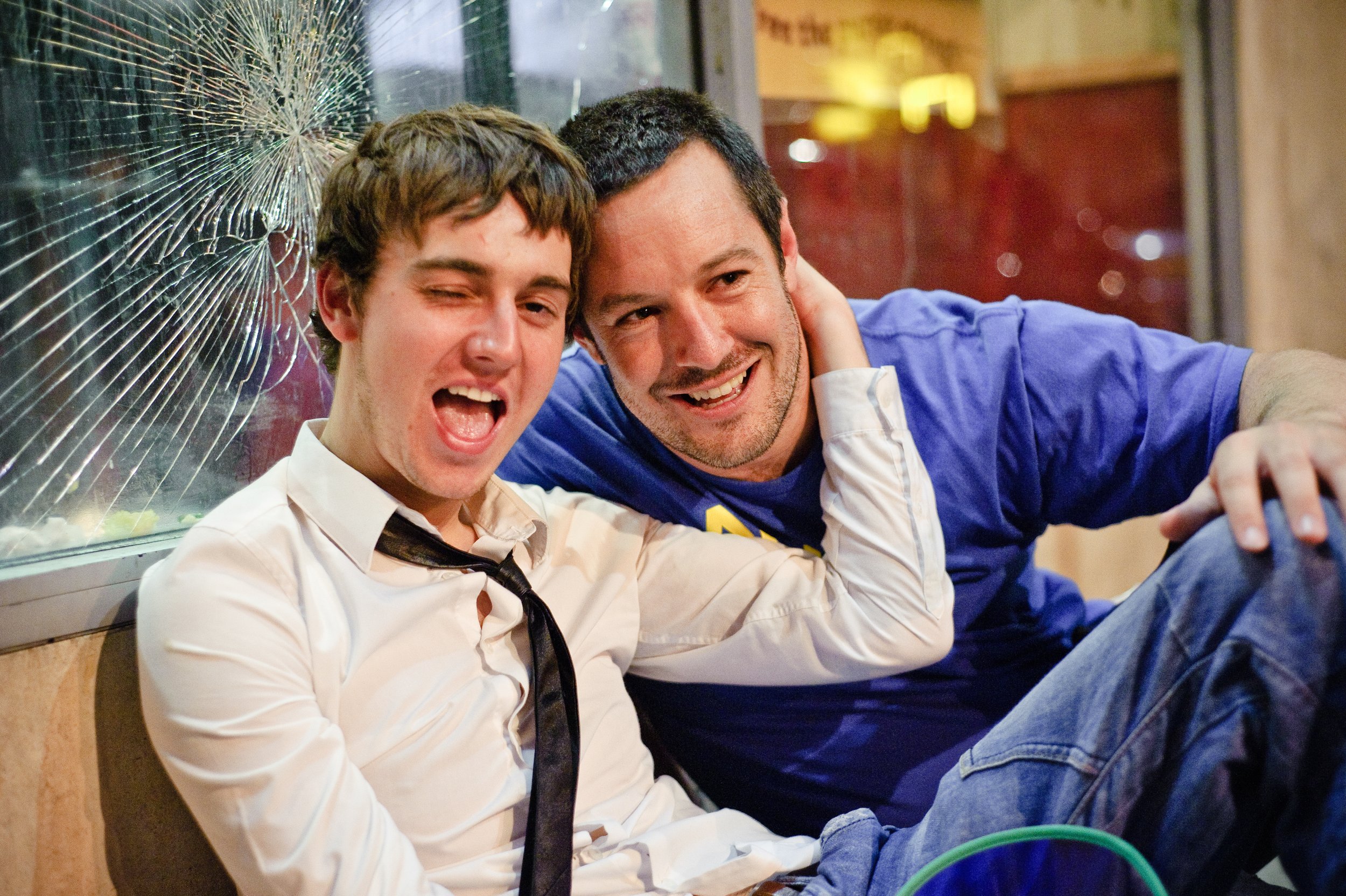 Two men sitting on the floor inside a building, with one man wearing a white shirt and black tie, and the other man wearing a blue long sleeve shirt. They are smiling and leaning against a window with a broken glass pane, with city lights in the back