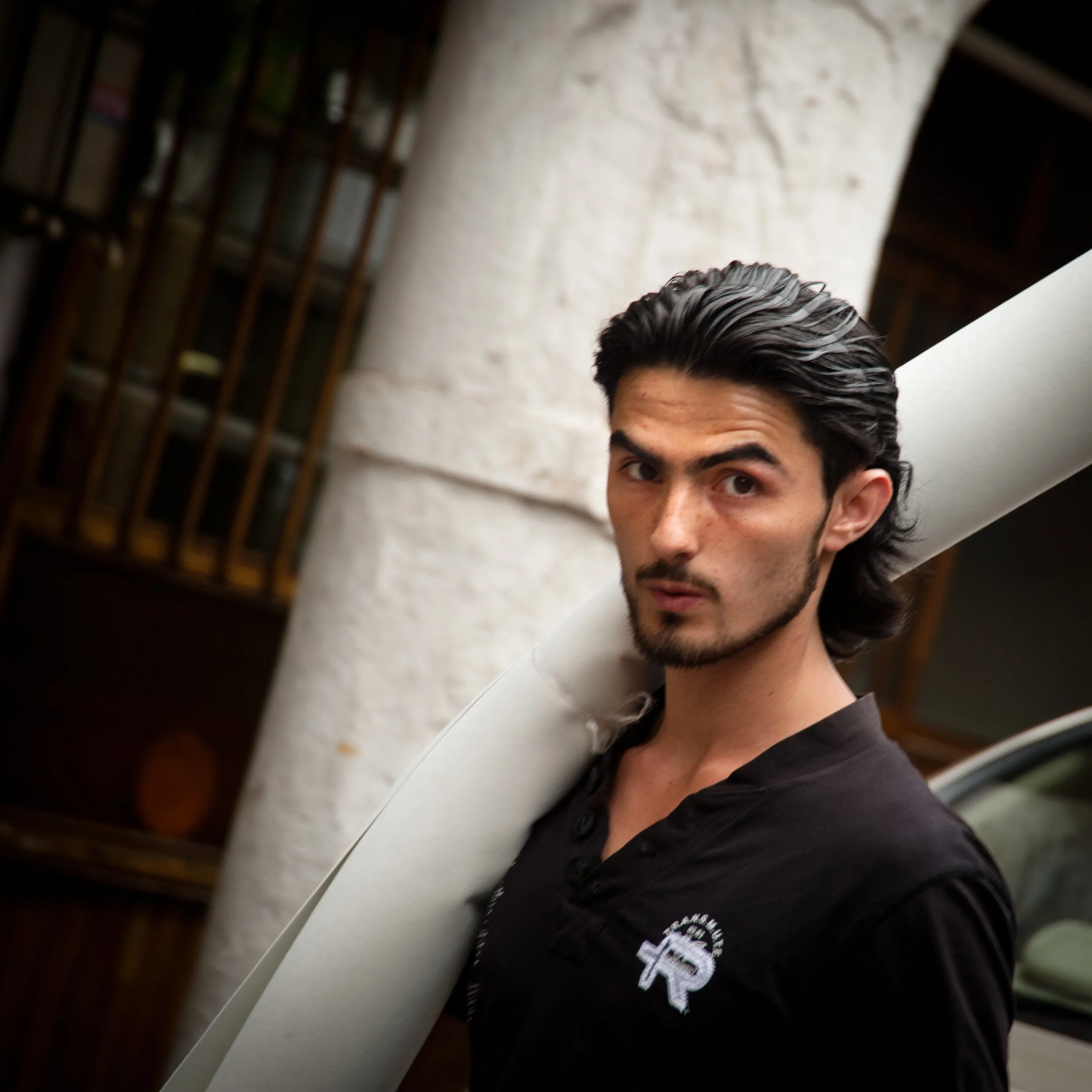 A man with dark, slicked-back hair and facial hair carrying a rolled-up white mat over his shoulder, wearing a black shirt with a white logo on it, standing outdoors near a white column and a wooden structure.