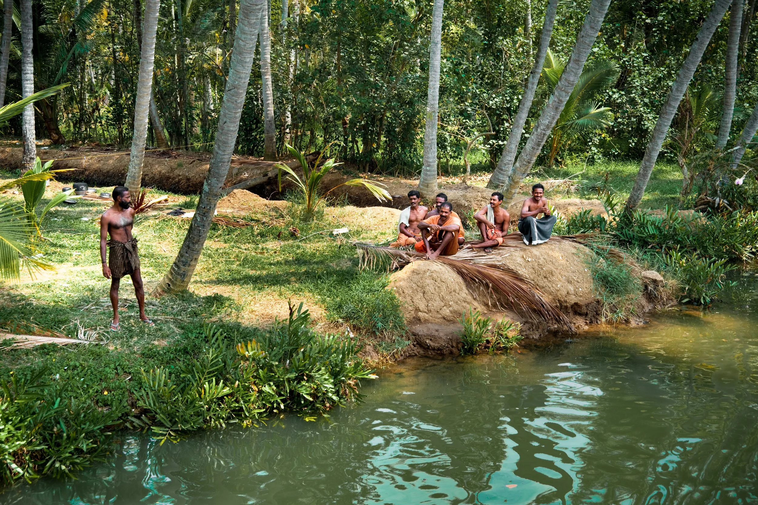 A group of five men sitting on a mound near a river in a lush, tropical forest, with one man standing nearby. The men are dressed in traditional clothing, and the scene appears to be a peaceful gathering in a natural setting.