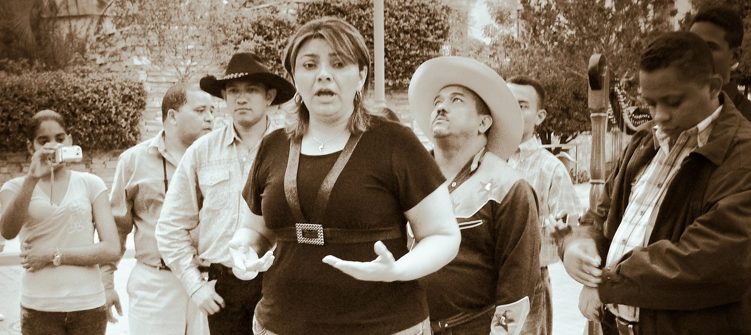 A group of people gathered outdoors, with a woman in the center speaking and gesturing with her hands, surrounded by men and women, some wearing cowboy hats and casual clothing, others taking photos or listening attentively.