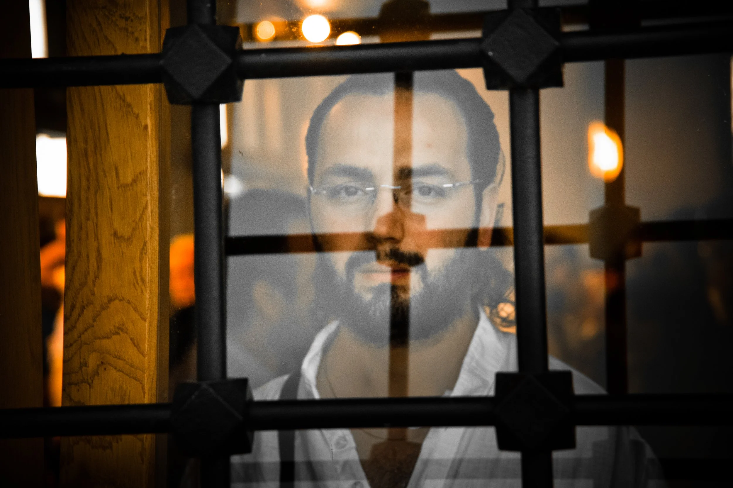 A reflection of a man with glasses and a beard, seen through black metal bars and a wooden frame.