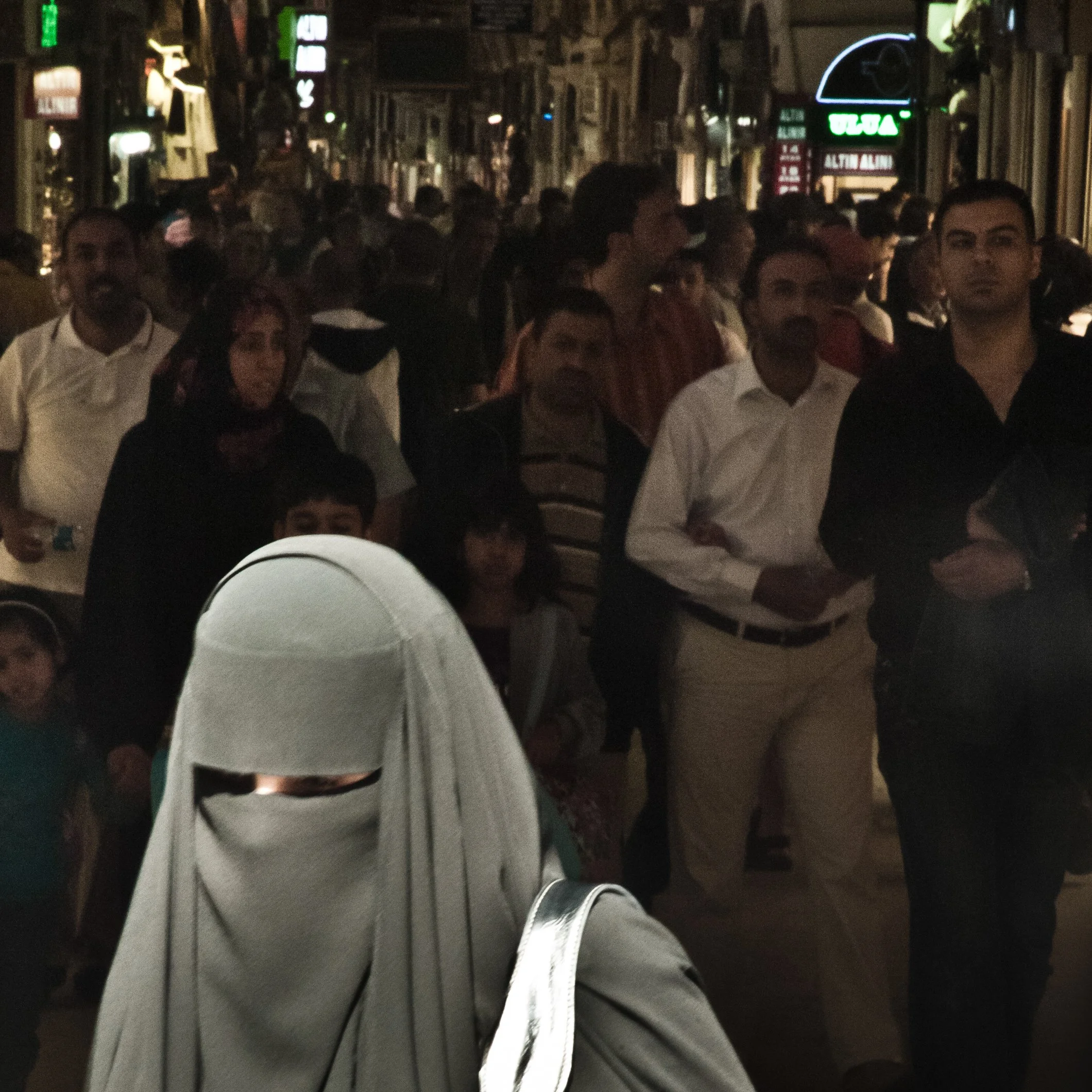 A crowded street scene at night with many people walking, including a woman wearing a white hijab in the foreground. Neon signs are visible in the background.