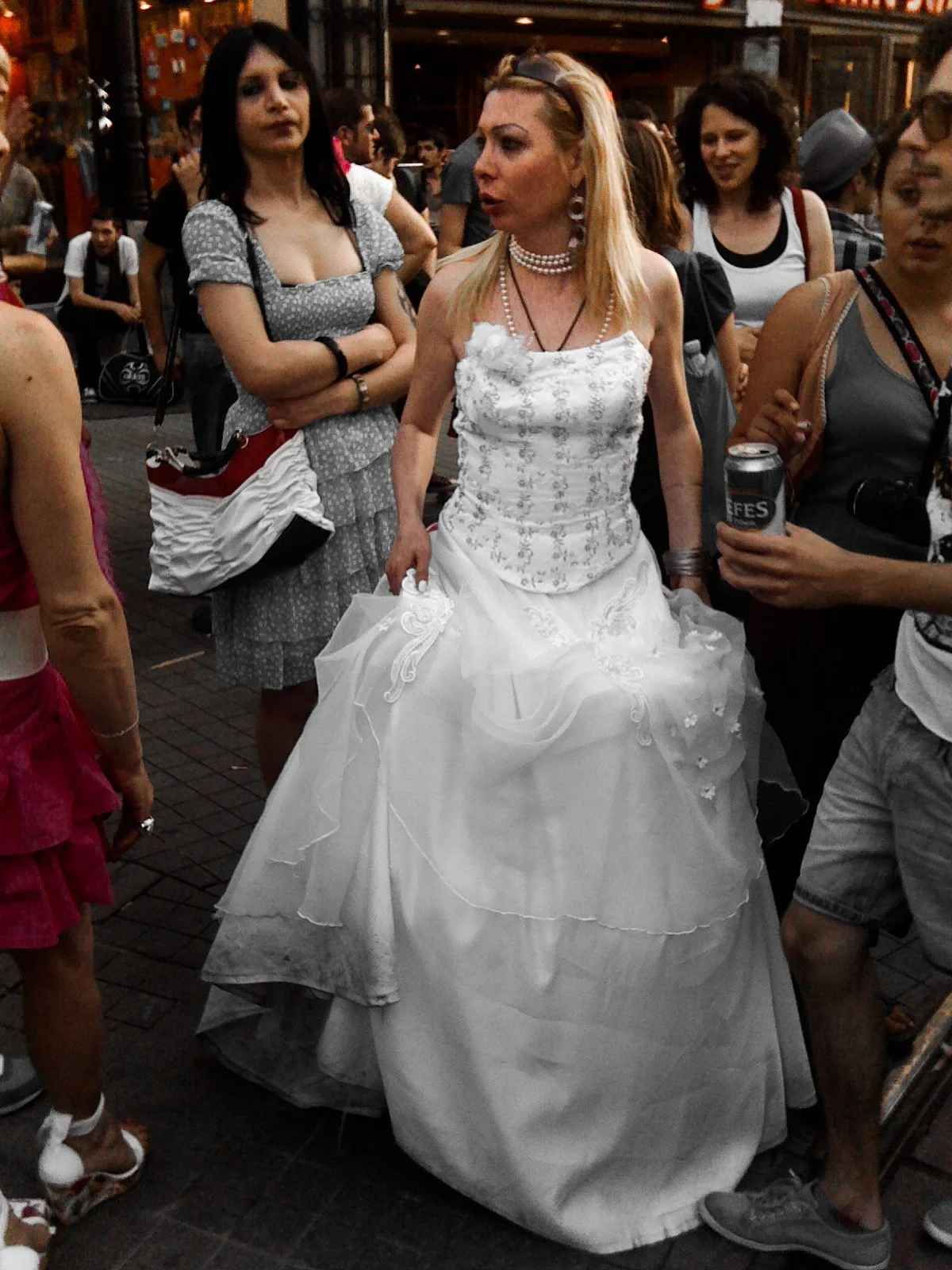 Woman wearing a white wedding dress in a crowded outdoor setting, surrounded by several people.