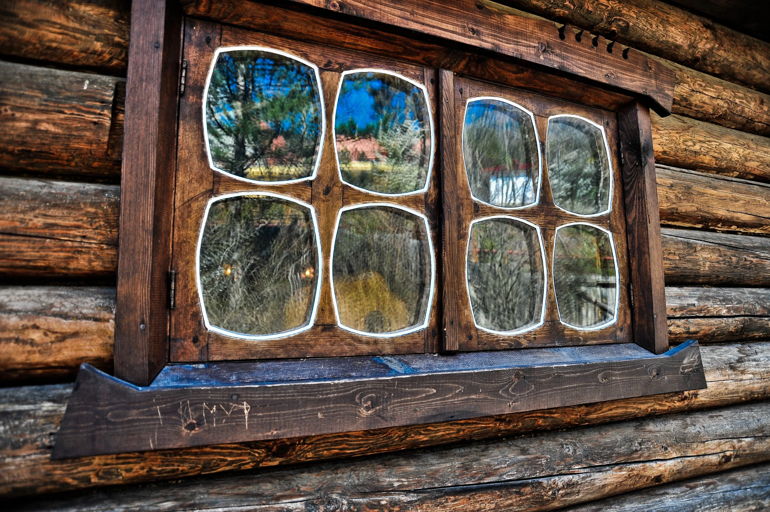 A wooden window with irregularly shaped glass panes on a log cabin wall, reflecting trees and a blue sky.