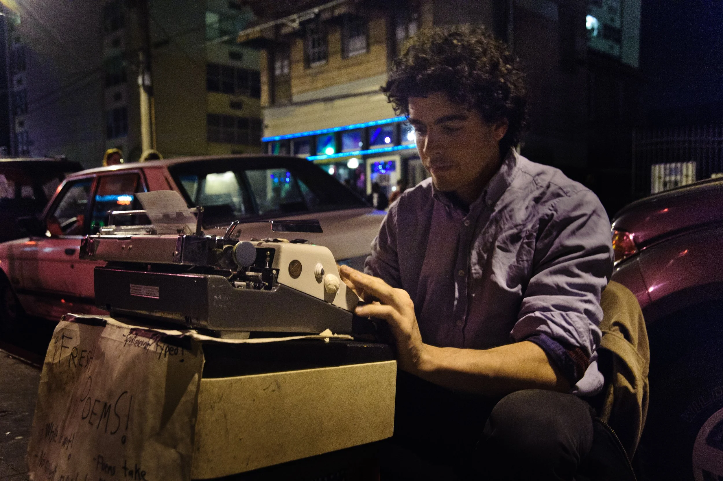 A man with curly hair sits on a city street at night, using a typewriter placed on a cardboard box, with cars and buildings illuminated by streetlights and neon lights in the background.