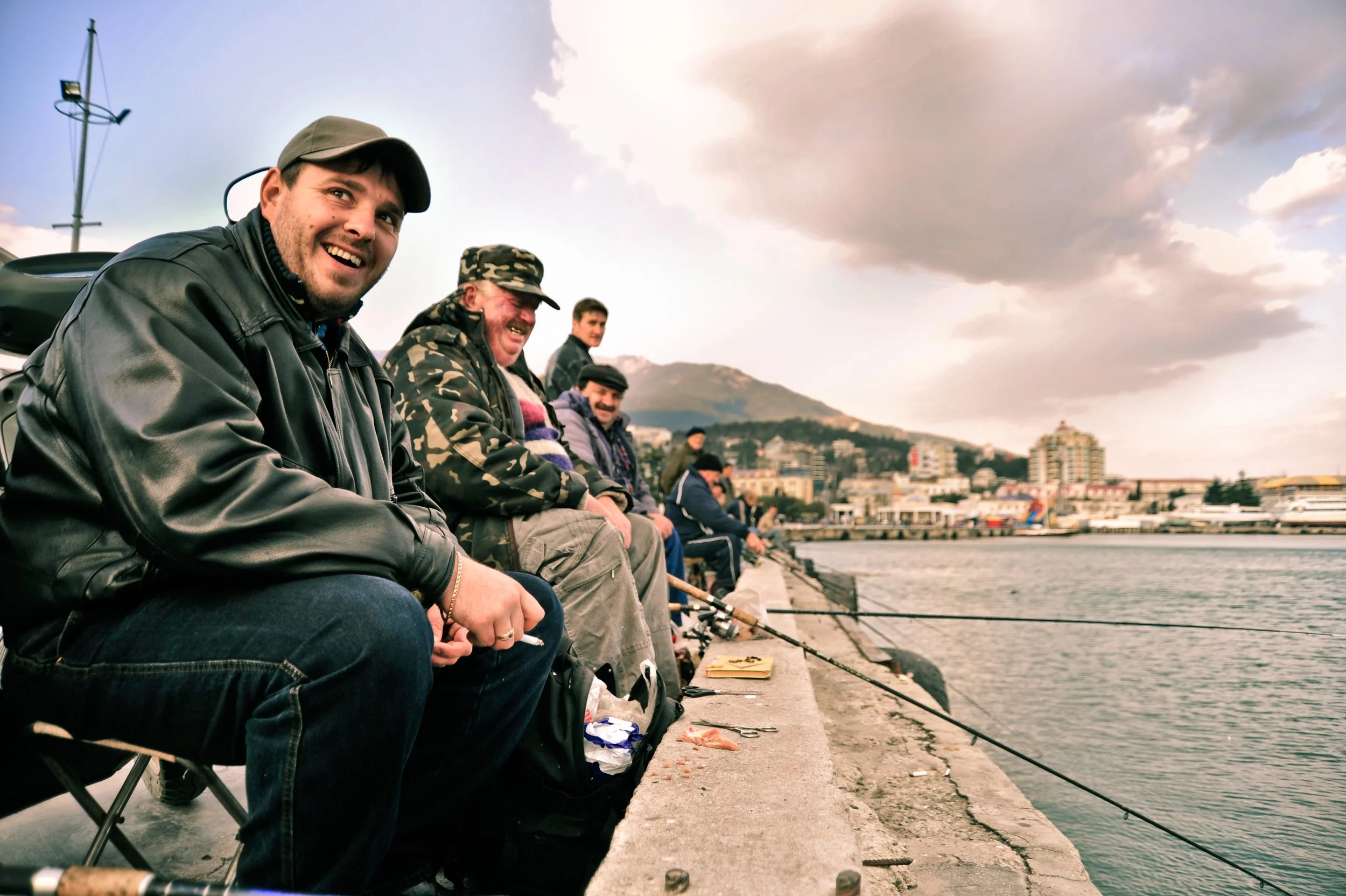 A group of men sitting on a harbor sidewalk fishing, with a cityscape and mountains in the background during the evening.