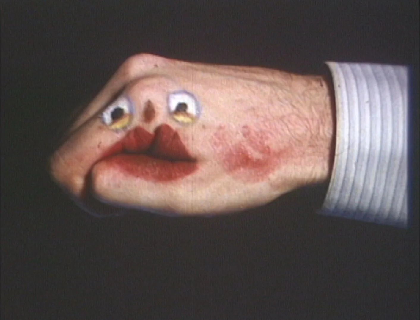 A strange and surreal image of a person's face with exaggerated features, including large eyes, red lips, and a nose painted on their cheek, blending exaggerated face features with a hand-placed painted face, set against a dark background.