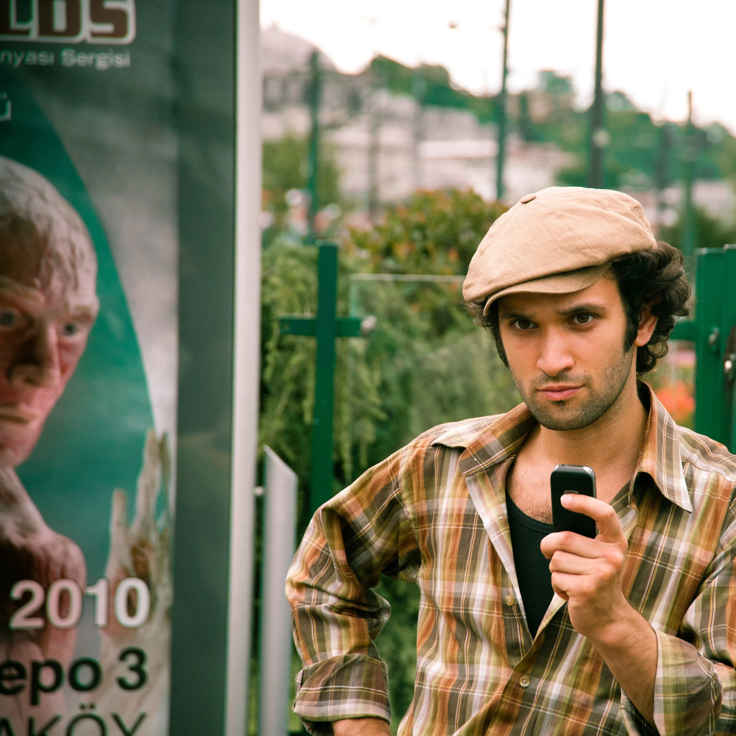 A man with curly dark hair, wearing a beige cap and a plaid shirt, holds a small black device and looks directly at the camera.