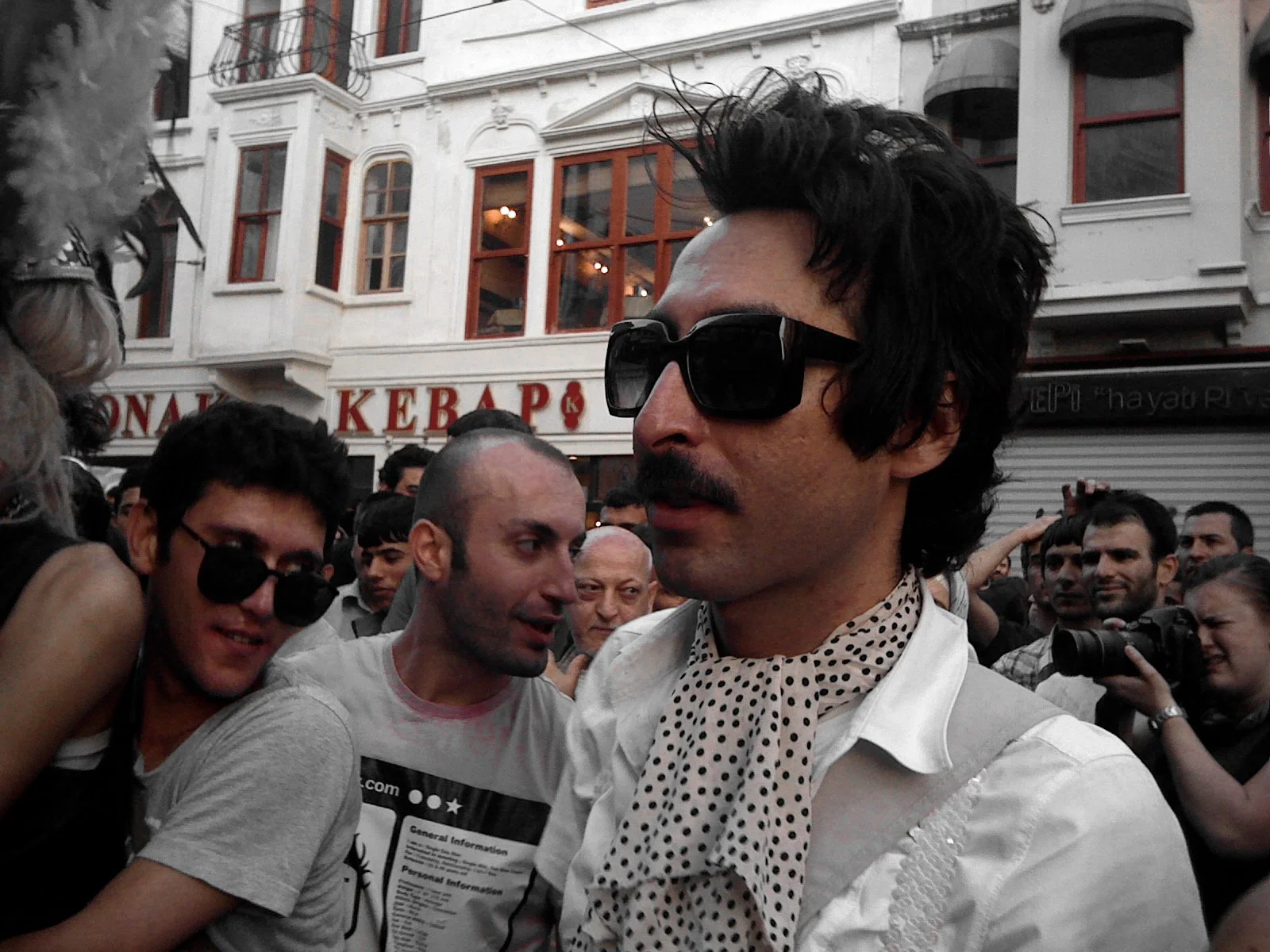 A man with dark hair, mustache, and sunglasses wearing a white shirt and polka dot scarf around his neck is surrounded by a crowd of people in an urban setting. The background shows a multi-story building with a sign in Turkish.