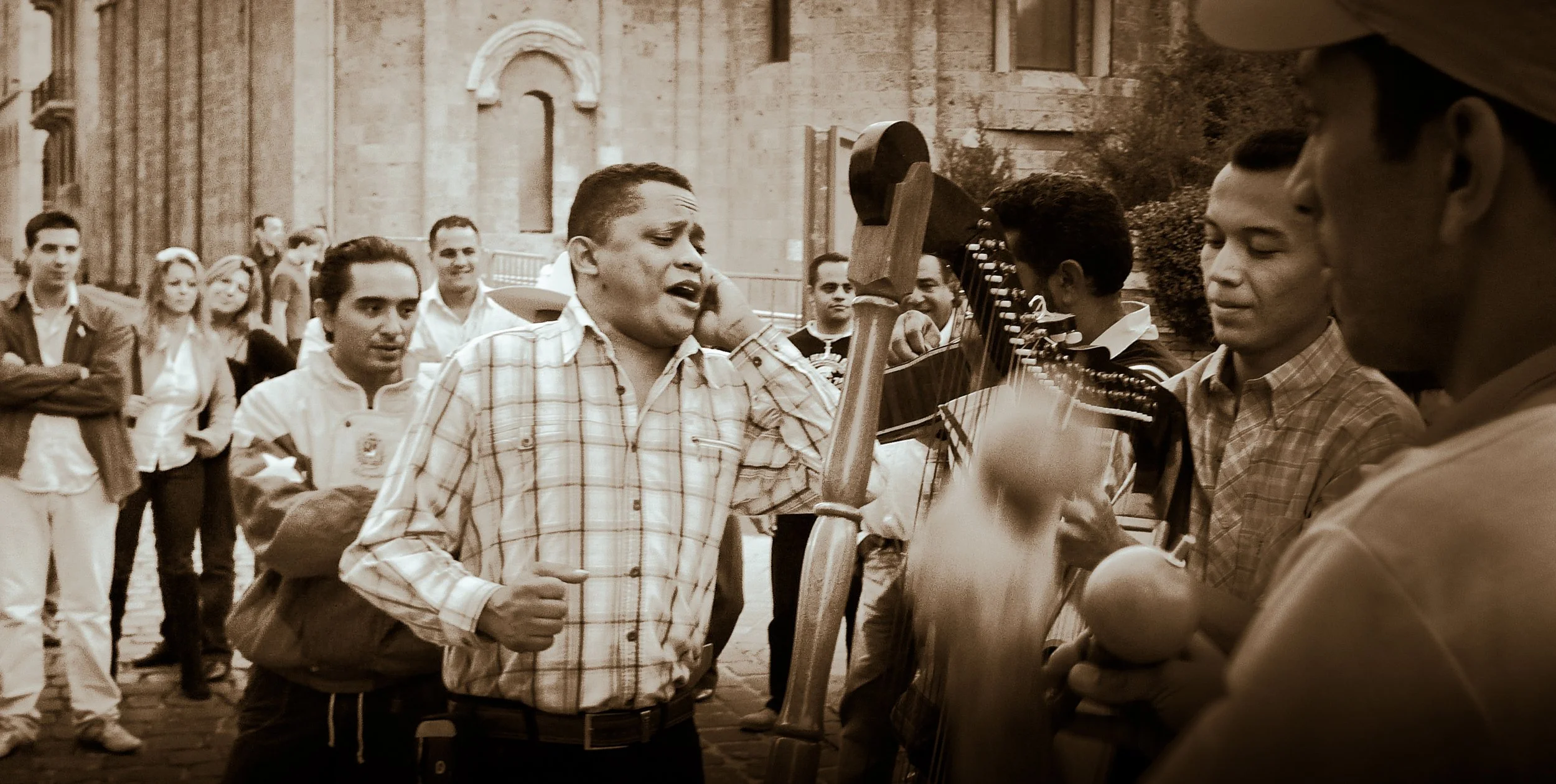 A group of people, including a man singing passionately, gathers outdoors with musicians holding guitars and a percussion instrument, in front of a historic building with stone architecture.