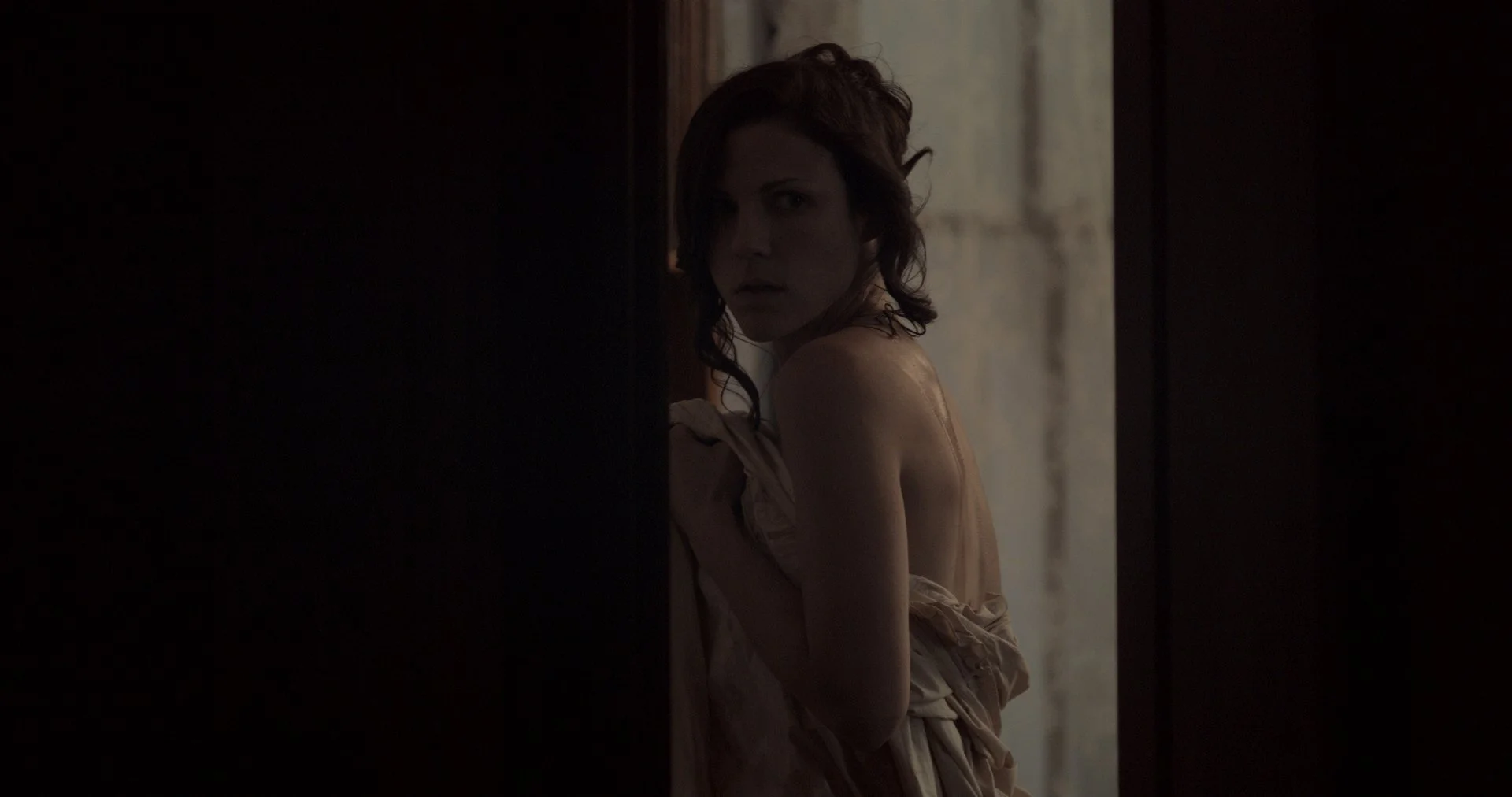 A woman with curly hair looks over her shoulder in a dimly lit room, partially obscured by a dark wall.