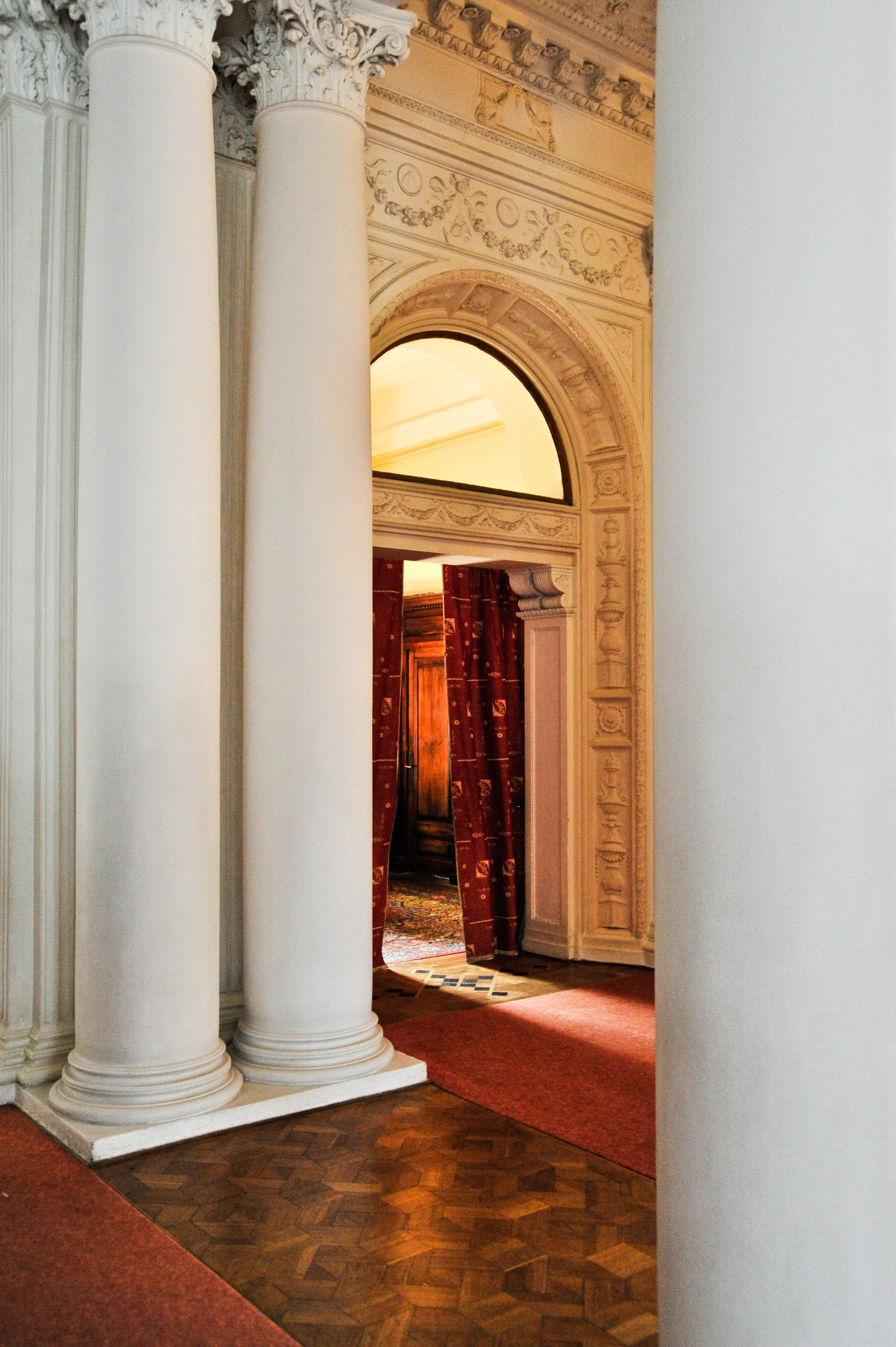 Elegant interior with large white columns, detailed architectural moldings, and a doorway framed with ornate trim revealing a room with red curtains and wooden furniture.