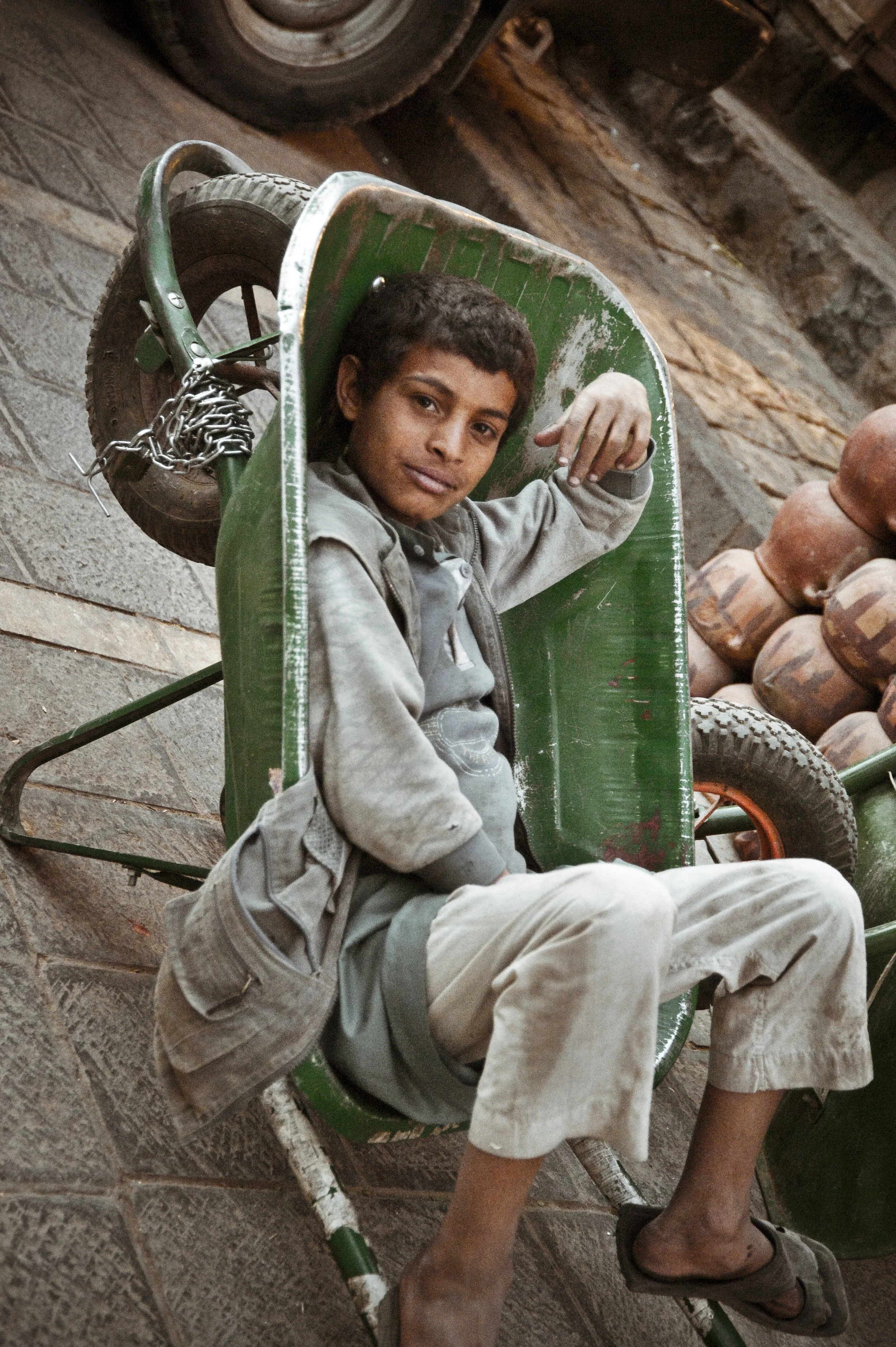 A young man sitting in a green wheelbarrow on a stone pavement, with pottery in the background.