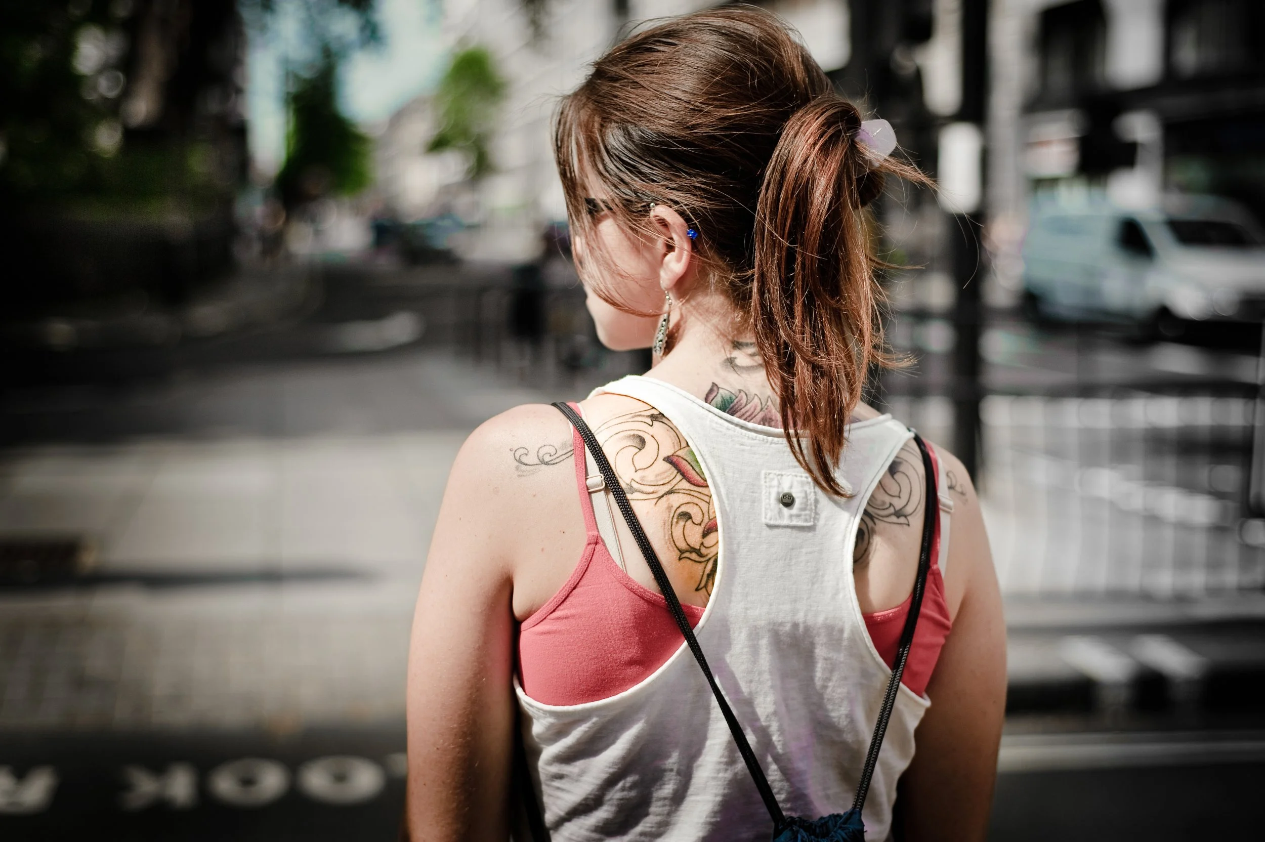 A woman with tattoos and brown hair tied in a ponytail, wearing a sleeveless shirt and earrings, seen from behind on a city street.