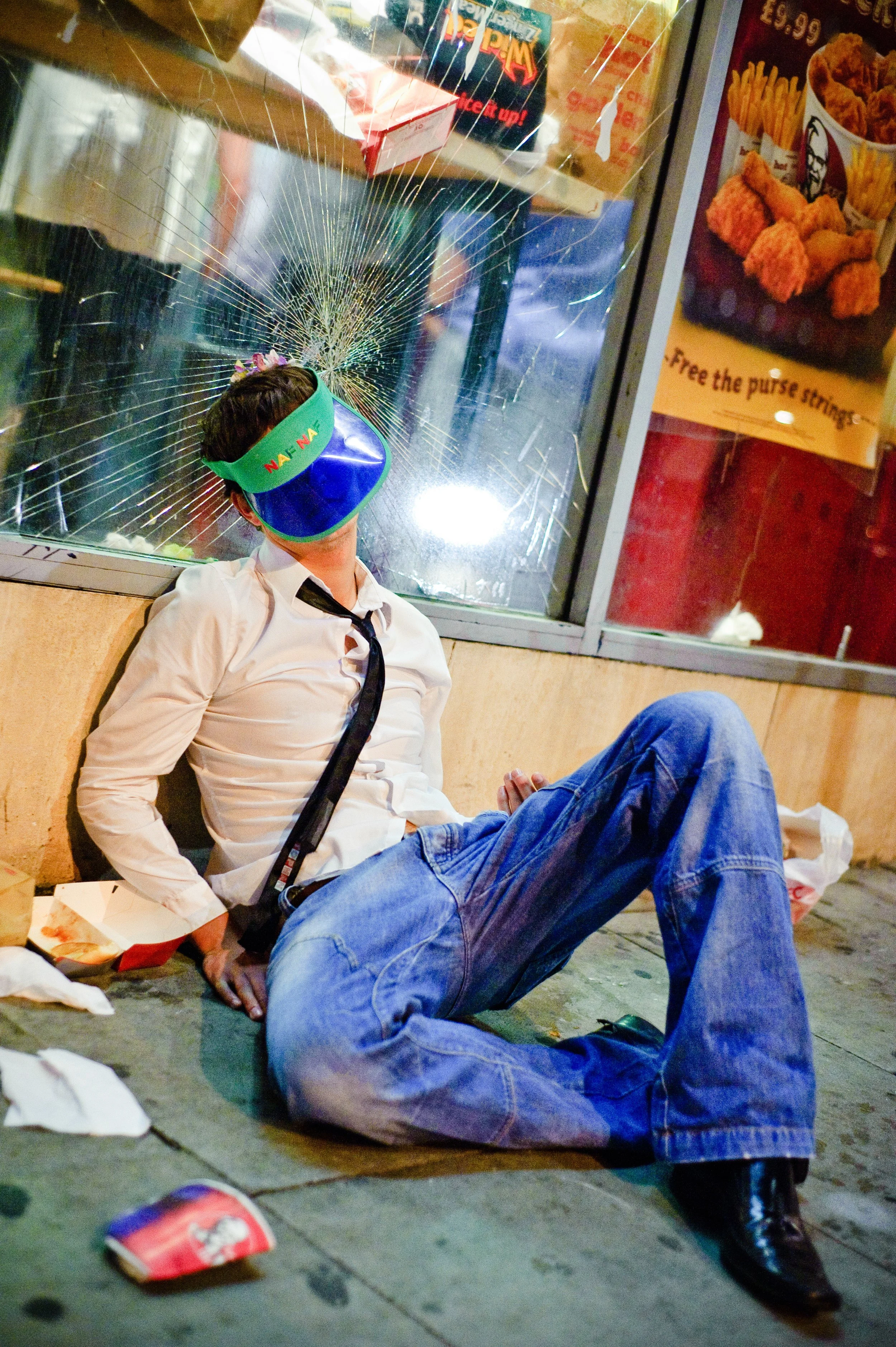 A man in a white shirt and tie is sitting on the sidewalk outside a store, leaning against a wall with his eyes covered by a colorful visor. The store window in the background shows a broken glass pane with a spider web pattern.