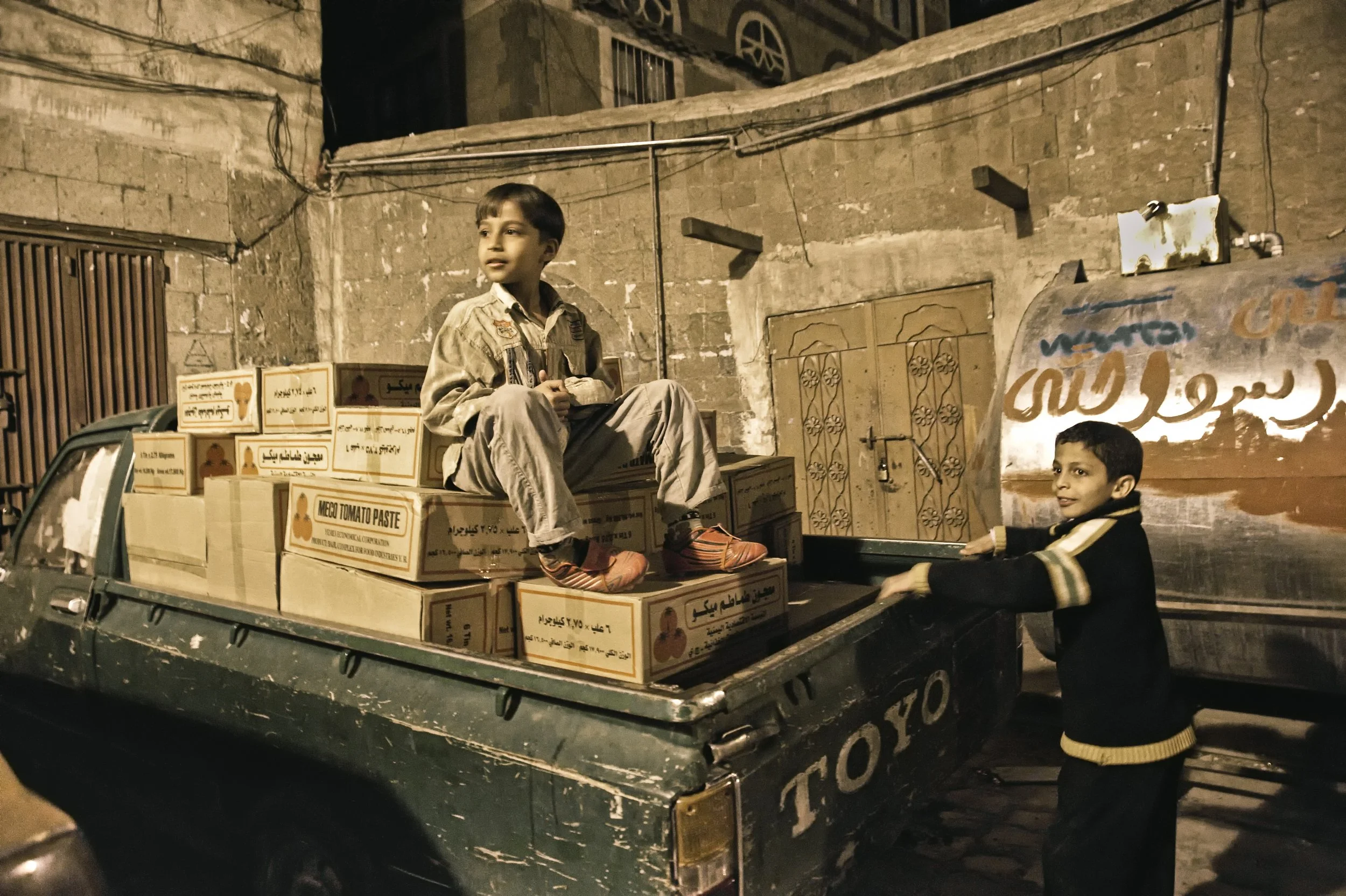 Two boys are loading boxes of tomato paste onto a small green truck in an alleyway at night. One boy is sitting on top of the boxes, while the other is standing and holding onto the truck.