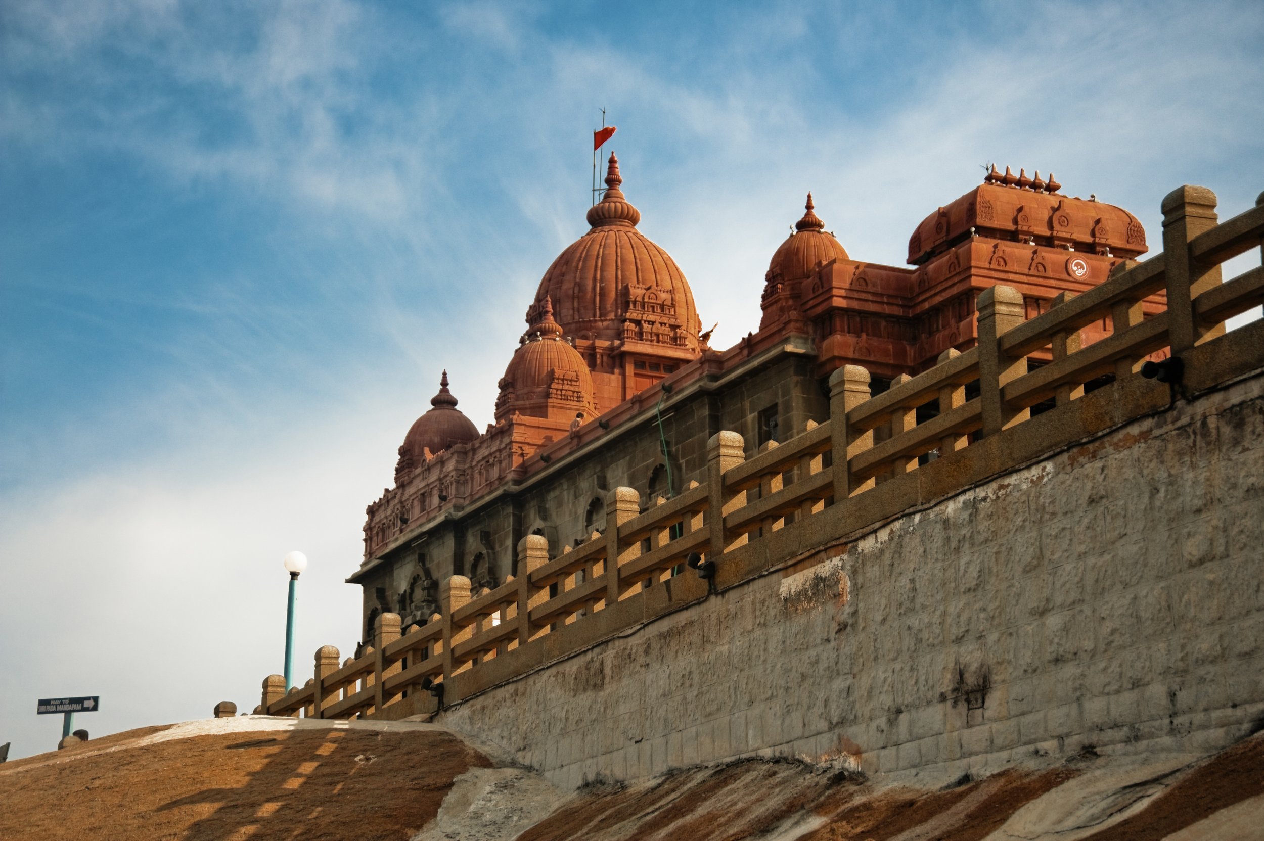 A historic building with red sandstone domes, possibly a temple or palace, seen from a low angle against a cloudy blue sky, with a stone wall and a fence in the foreground.
