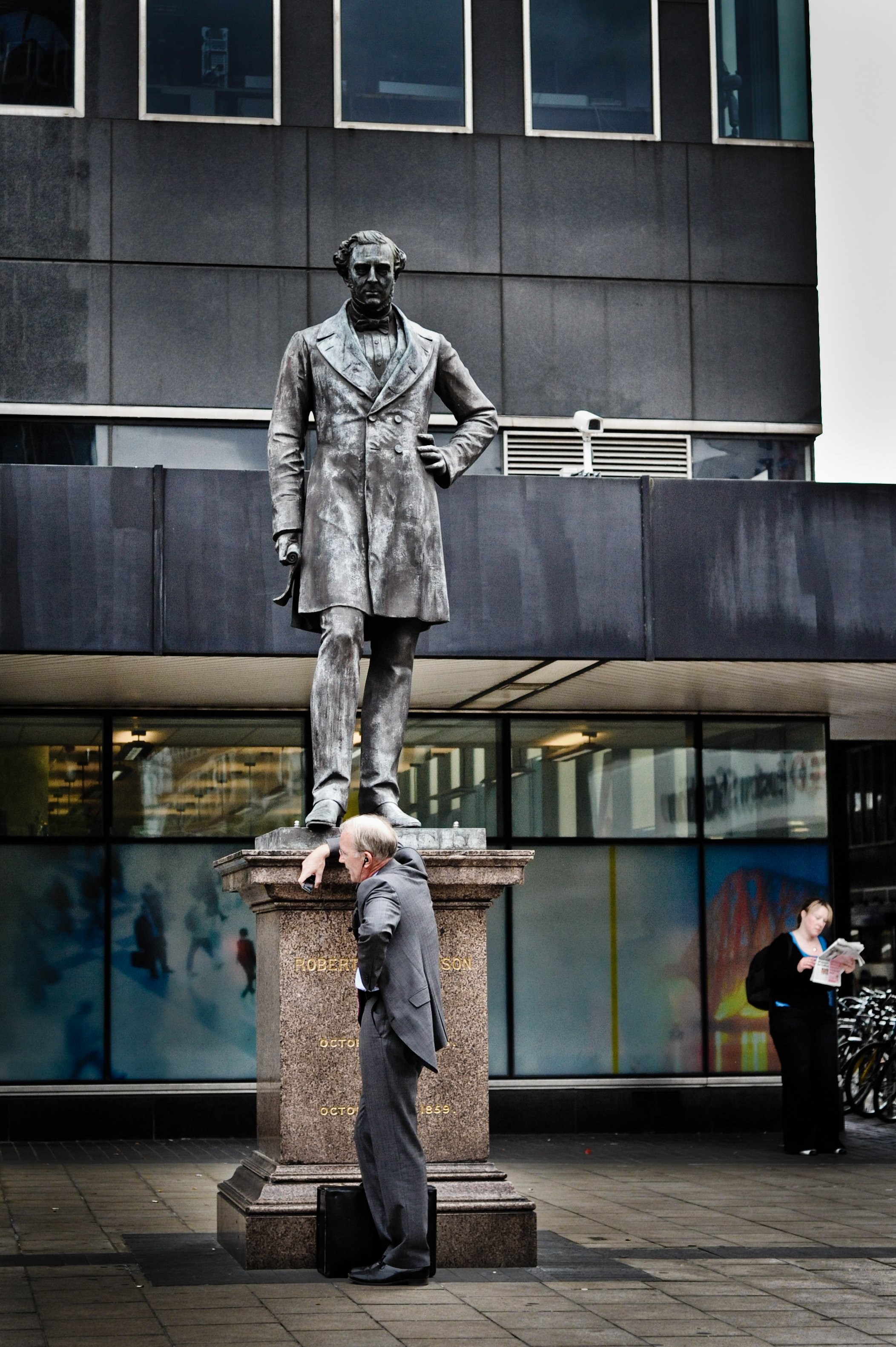 Man leaning on a statue of Robert E. Lee in an urban setting, with a woman reading a newspaper in the background.