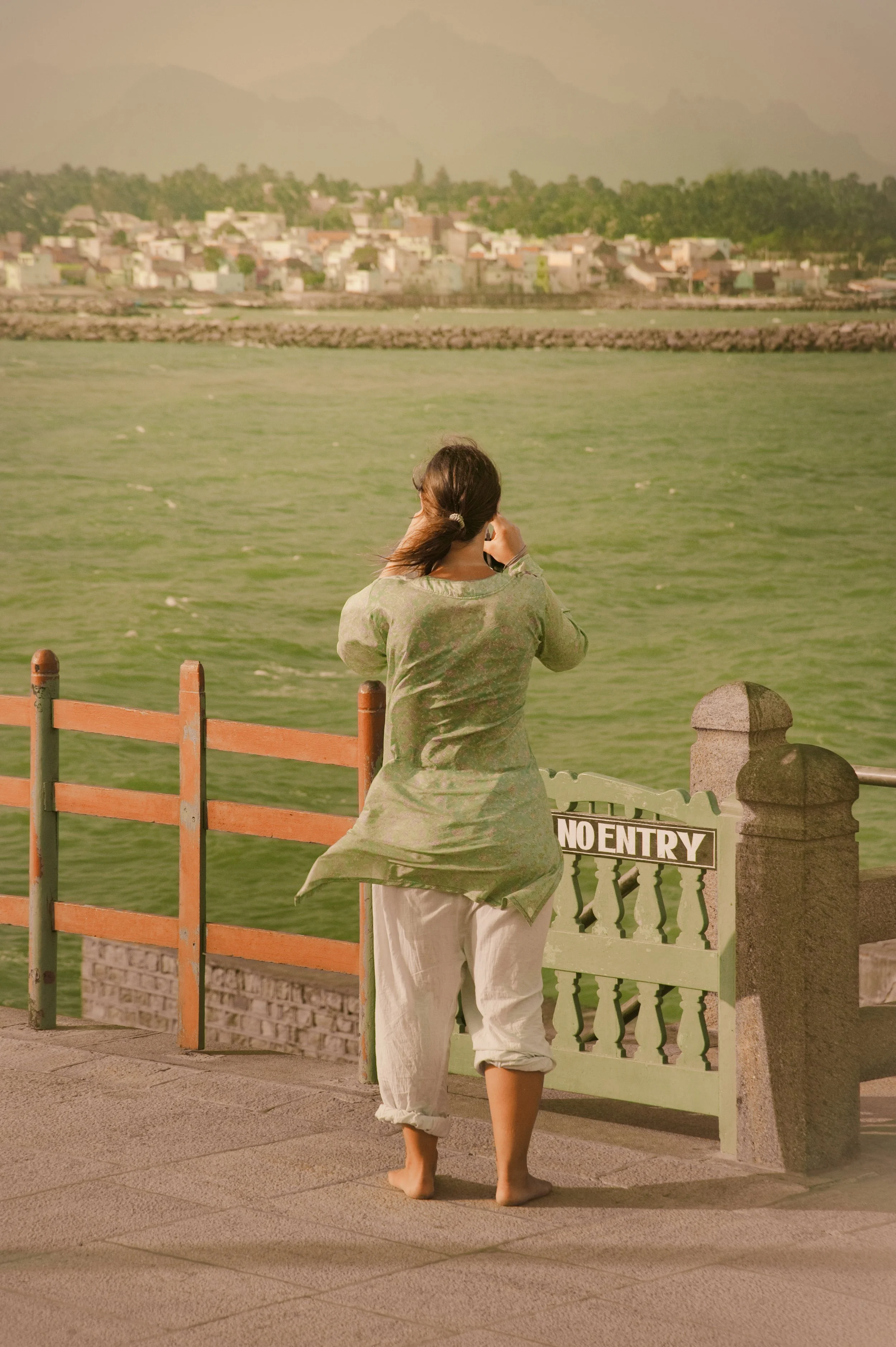 A woman standing barefoot on a concrete walkway by a body of water, talking on her cellphone, with her back to the camera. She is wearing a light green kurta and white pants. There is a small green gate with a 'No Entry' sign blocking access to the w