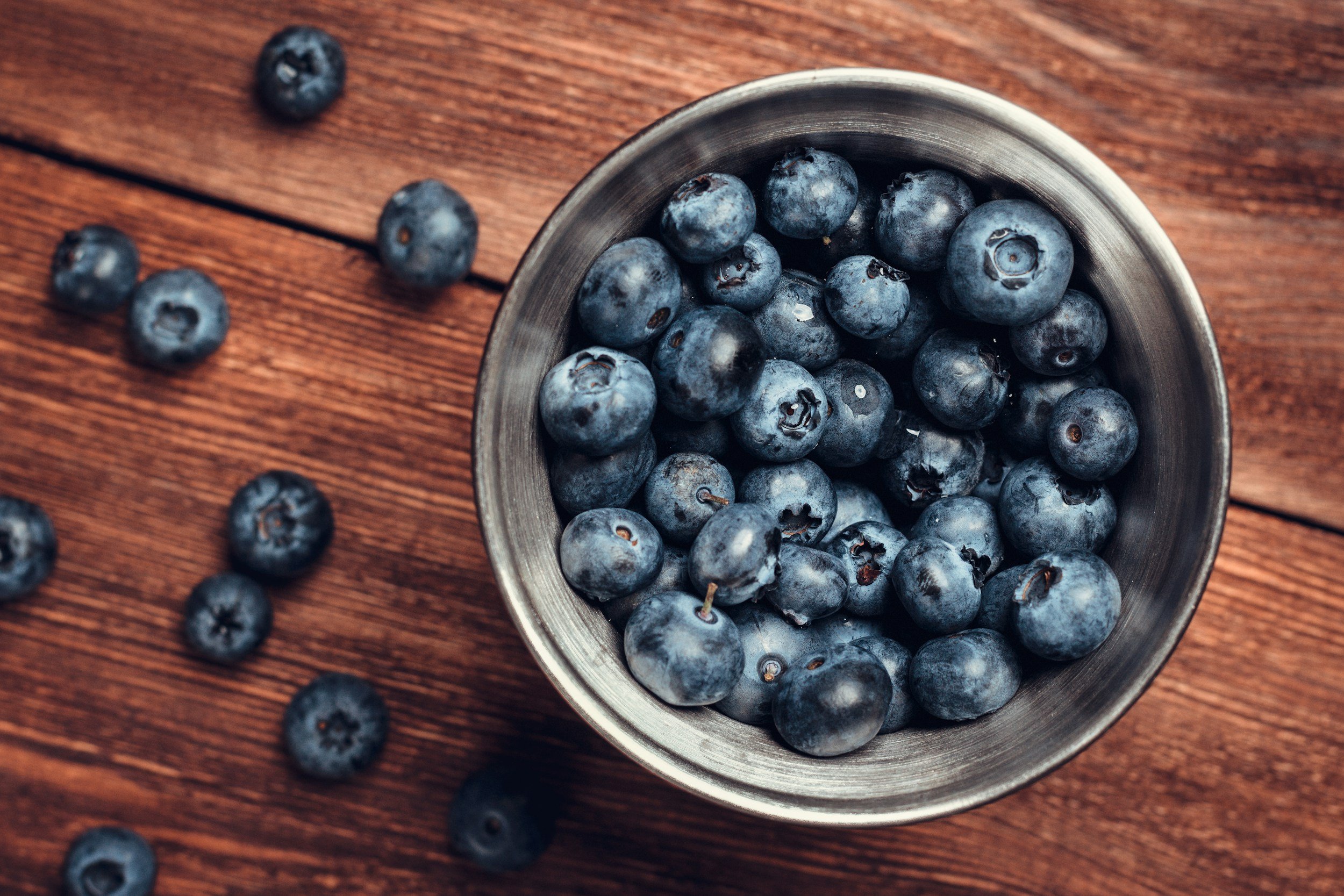 A metal bowl filled with fresh blueberries on a wooden surface with a few blueberries scattered around.