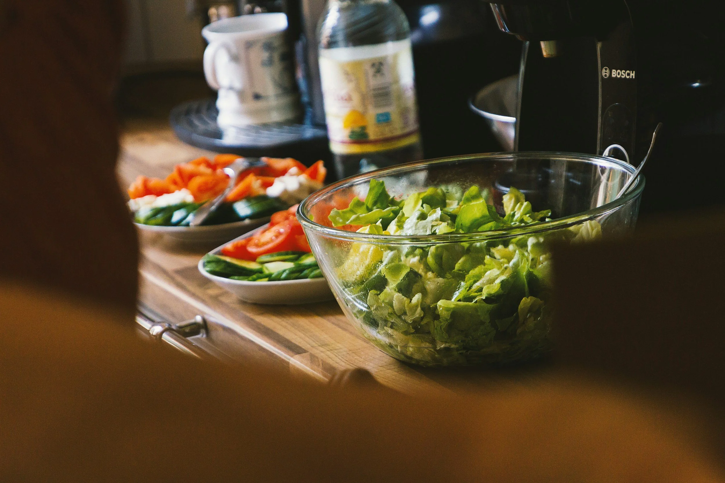 Bowl of chopped lettuce and plates of sliced cucumbers, tomatoes, and carrots on a wooden kitchen counter, with a coffee maker and bottles in the background.