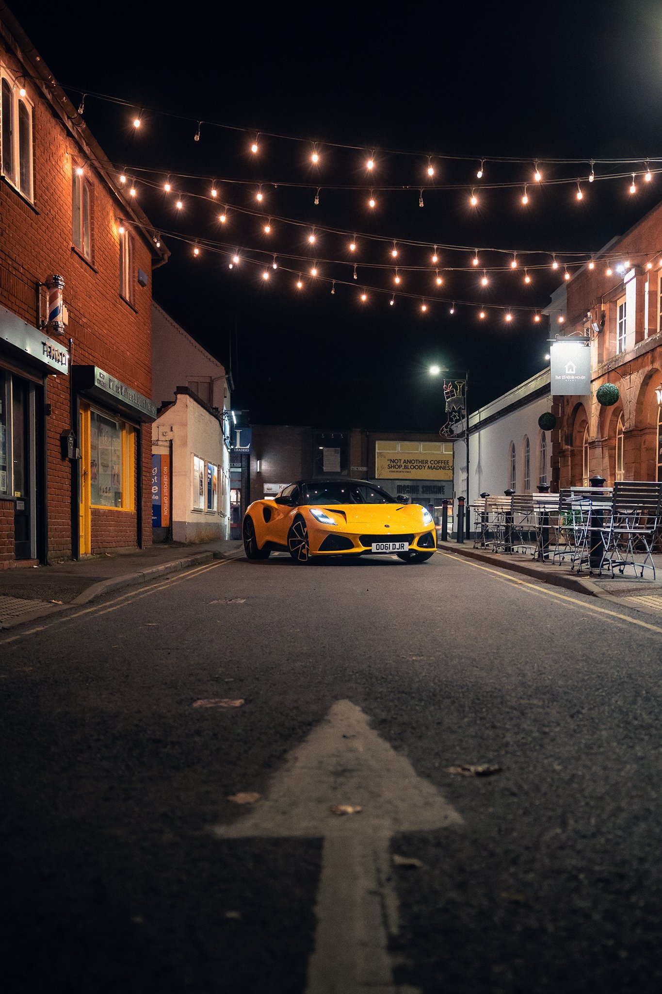 A yellow Lotus Emira posed outside of a restaurant on a street at night