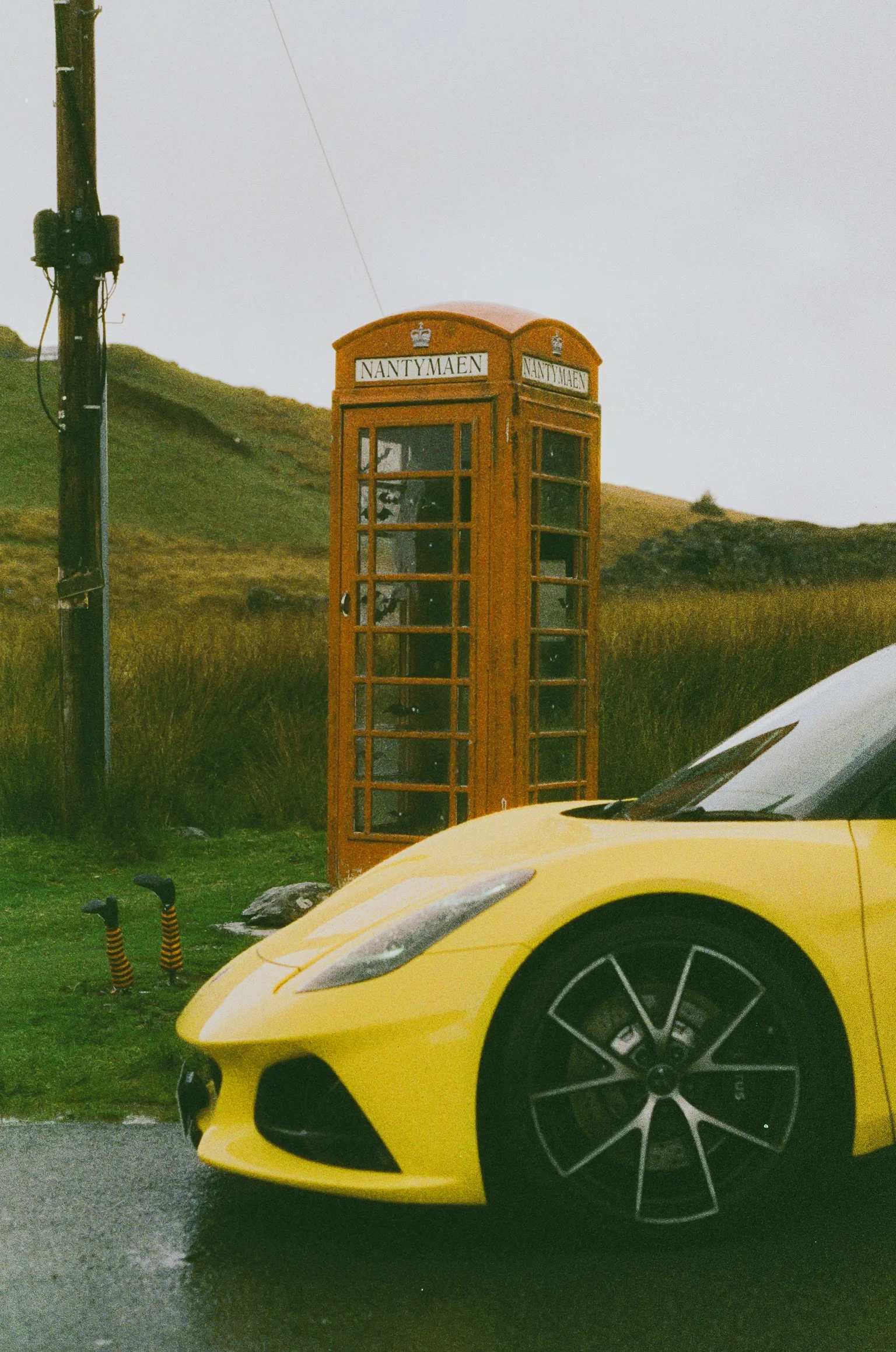 A yellow sports car parked near a red British phone booth with a grassy field and hills in the background.