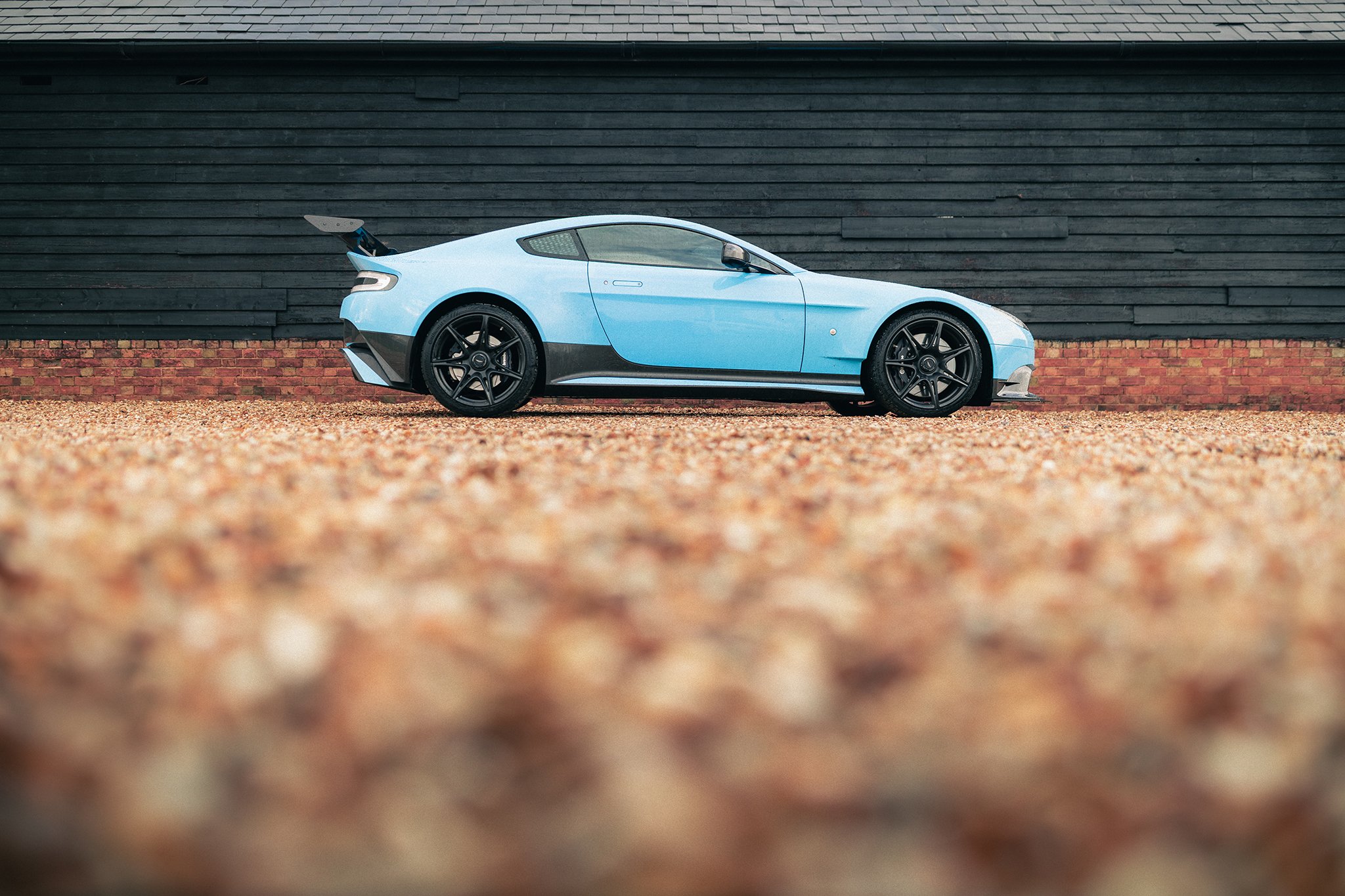 A Fugplatz Blue Aston Martin Vantage GT8 against a brick and wood panelled wall backdrop