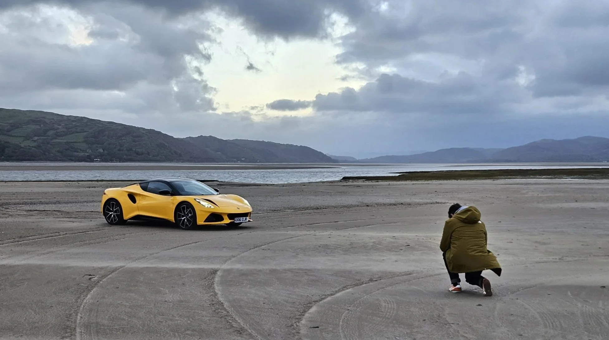 A yellow sports car parked on a sandy beach near water, with a person kneeling down taking a photo of the car, under a cloudy sky with distant mountains in the background.
