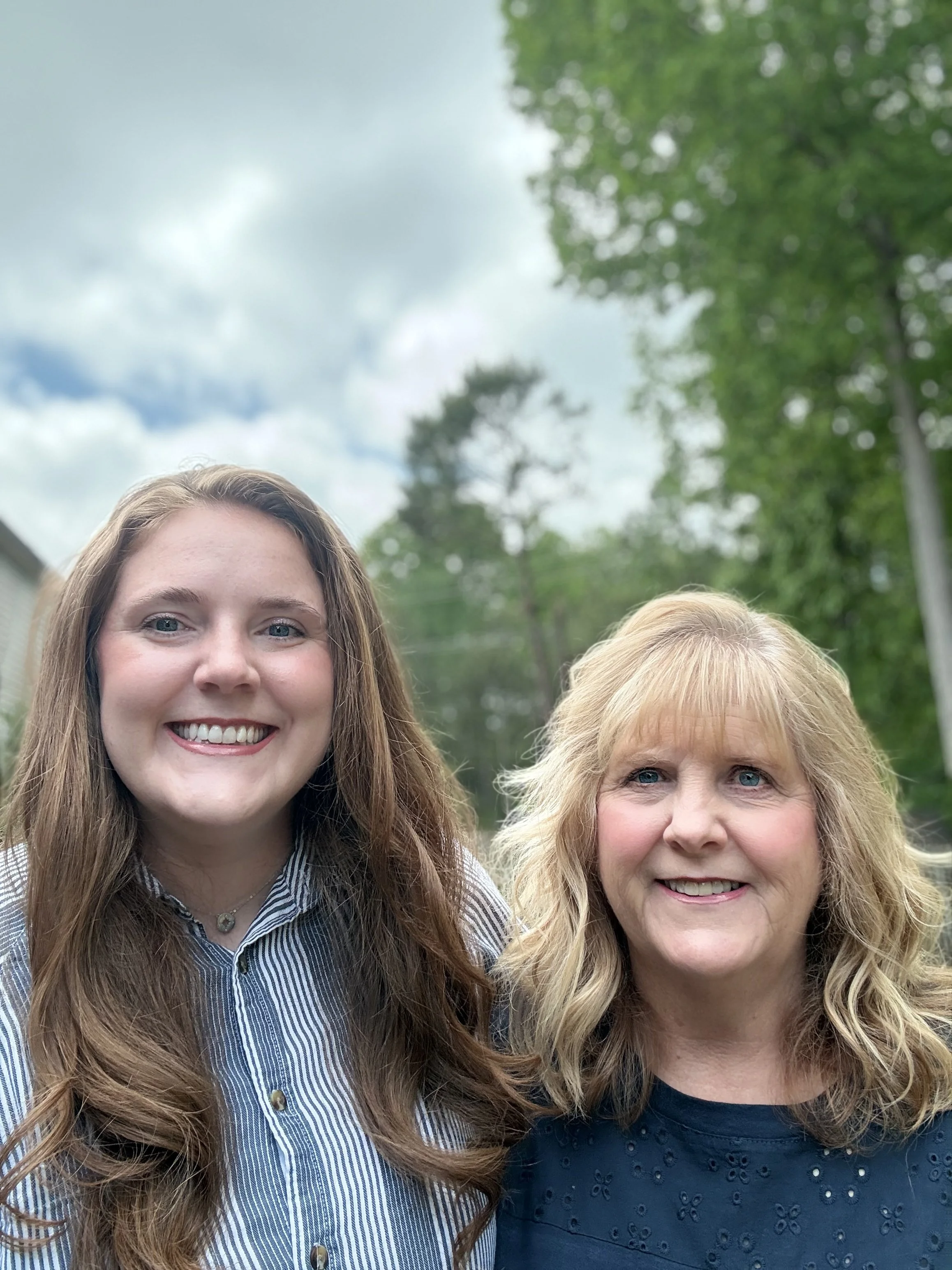 Two women with long hair smiling outdoors with a green leafy background.