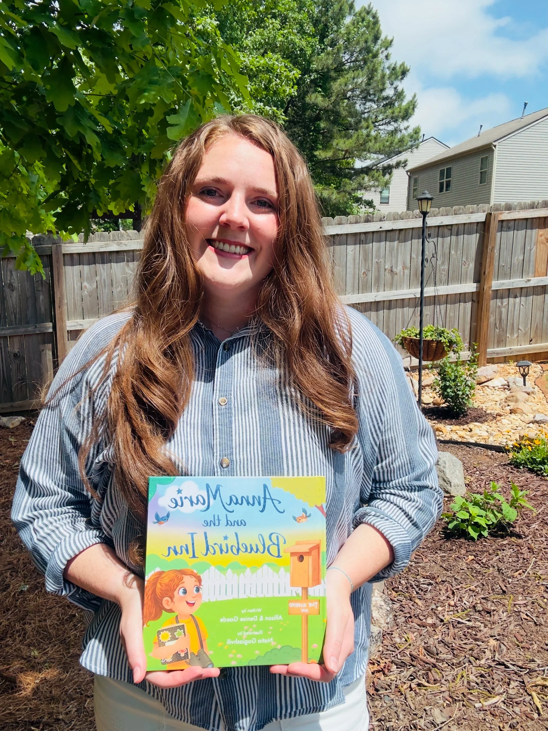 A young woman with long, wavy brown hair smiling outdoors in a backyard, holding a colorful children's book about planting a backyard garden.