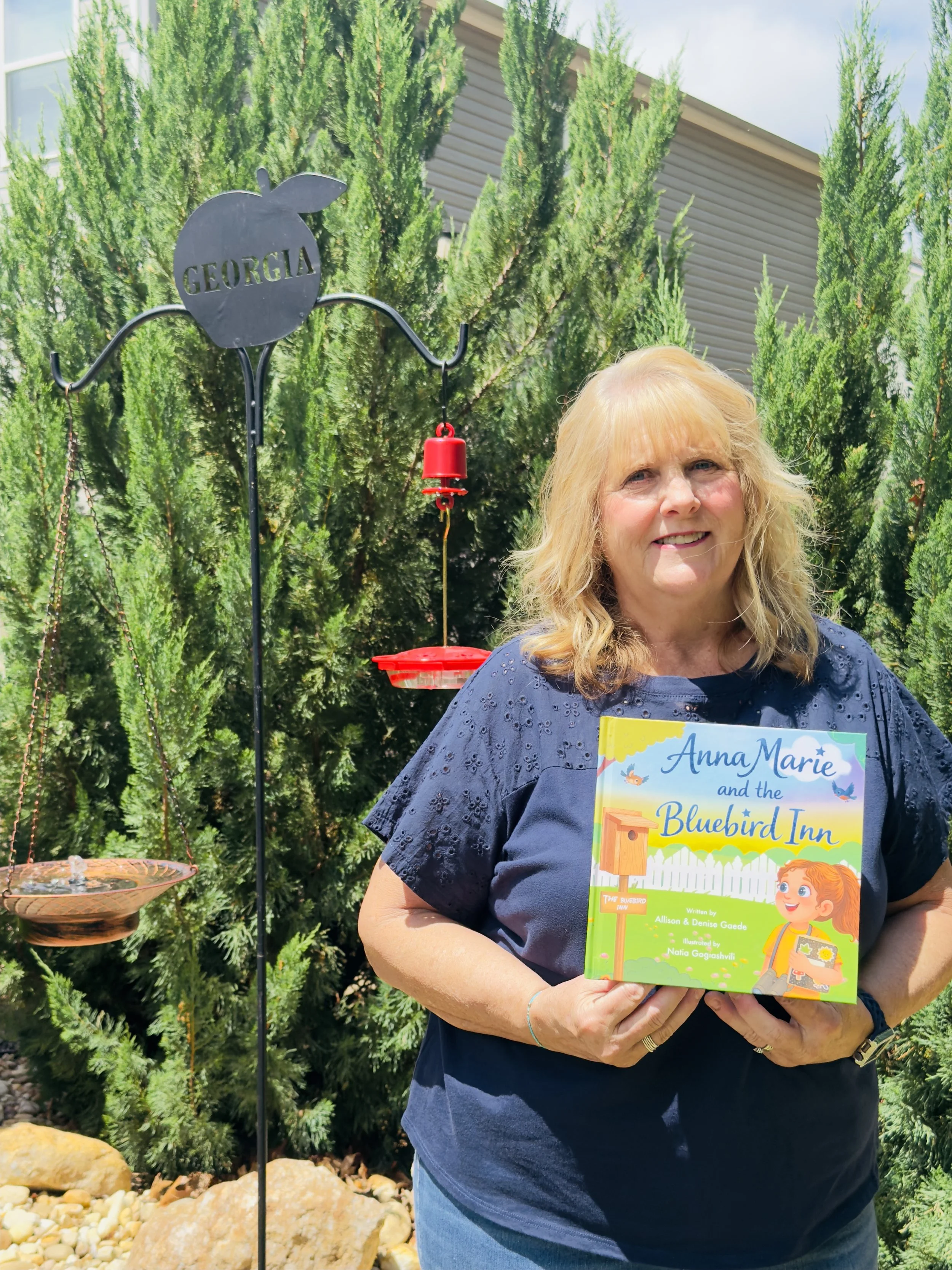 A woman with blonde hair holding a colorful children's book titled 'Anna Marie and the Bluebird Inn' outdoors, with green bushes and a house in the background, and a metal plant stand with a red bell hanging on it.